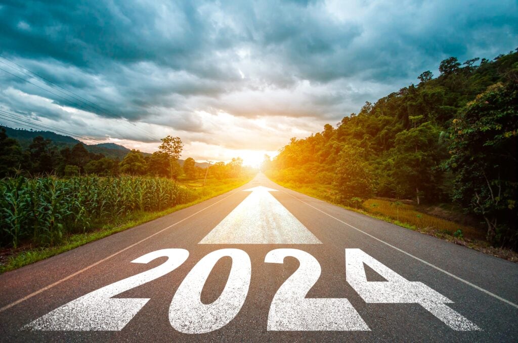 A straight road with "2024" and an arrow painted on it, surrounded by greenery under a cloudy sky with sunlight in the distance.
