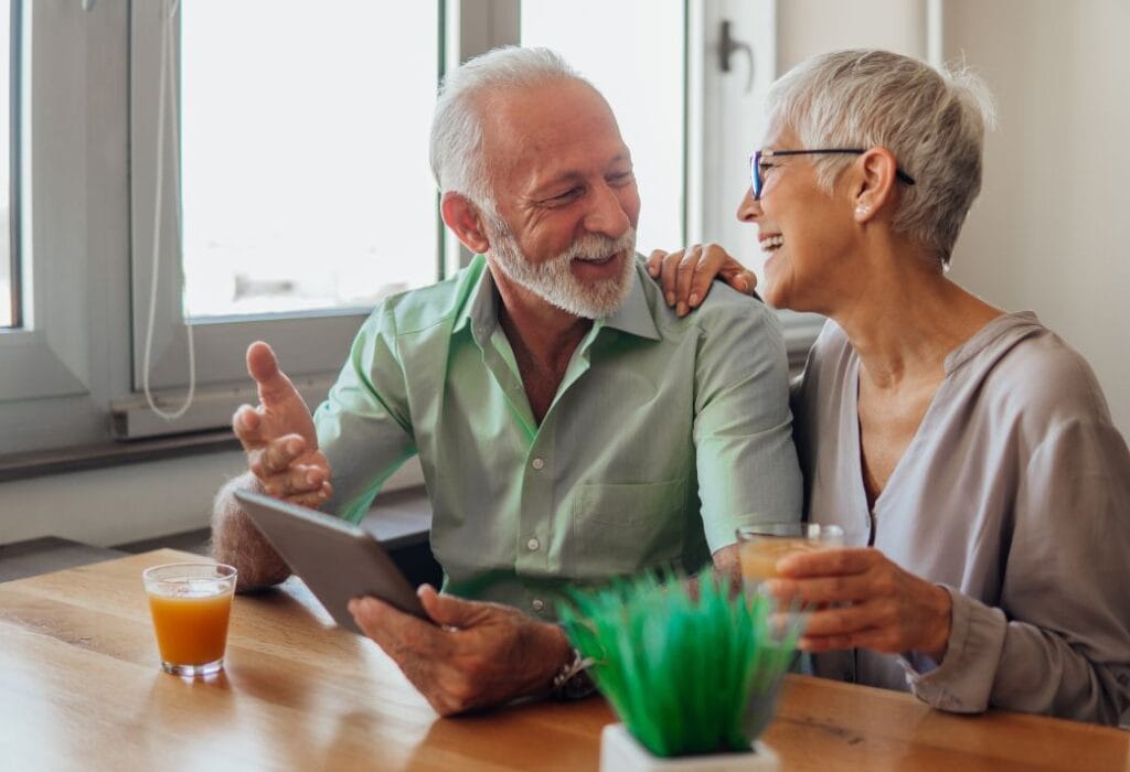 An elderly couple sits at a table with drinks, smiling and holding a tablet.