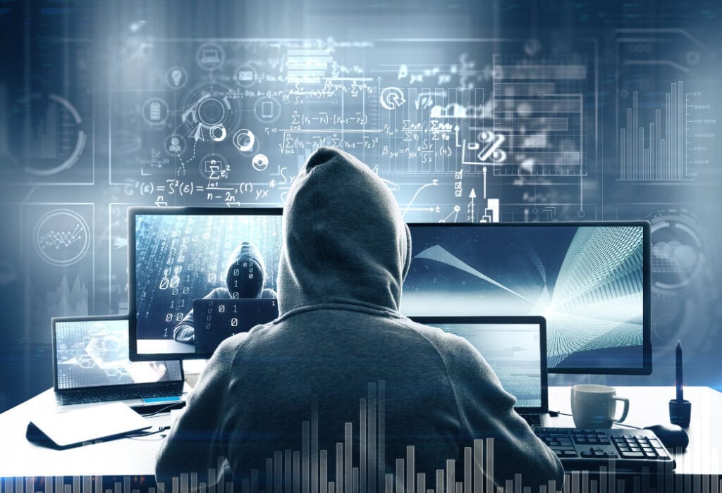A person in a hoodie works at a computer desk, surrounded by multiple screens displaying data and graphs.