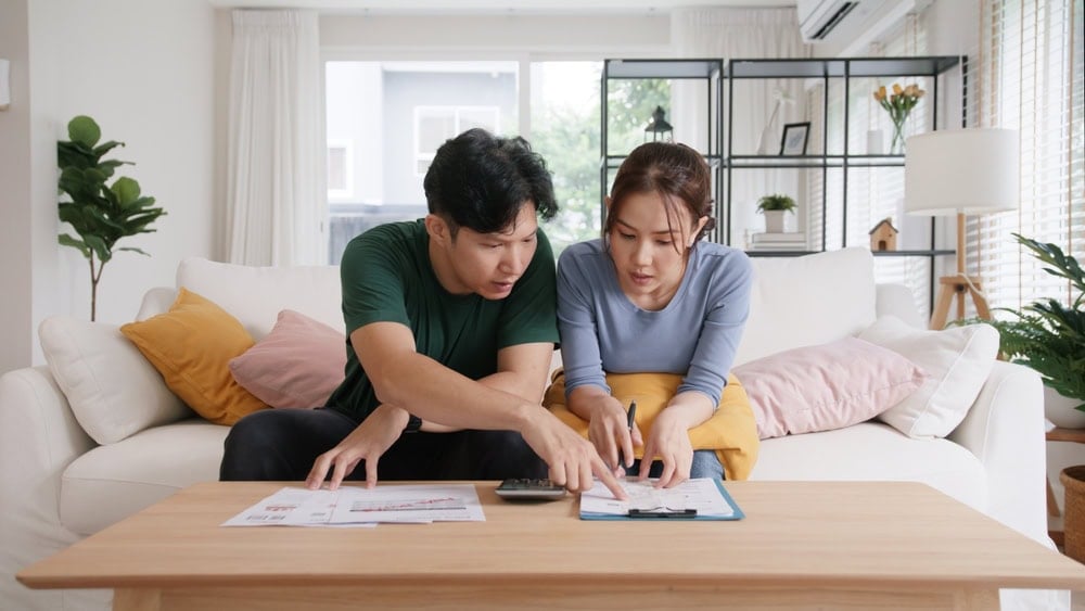 A couple sits on a couch, looking at documents on a coffee table, with a calculator in use. They seem focused and engaged in discussion.