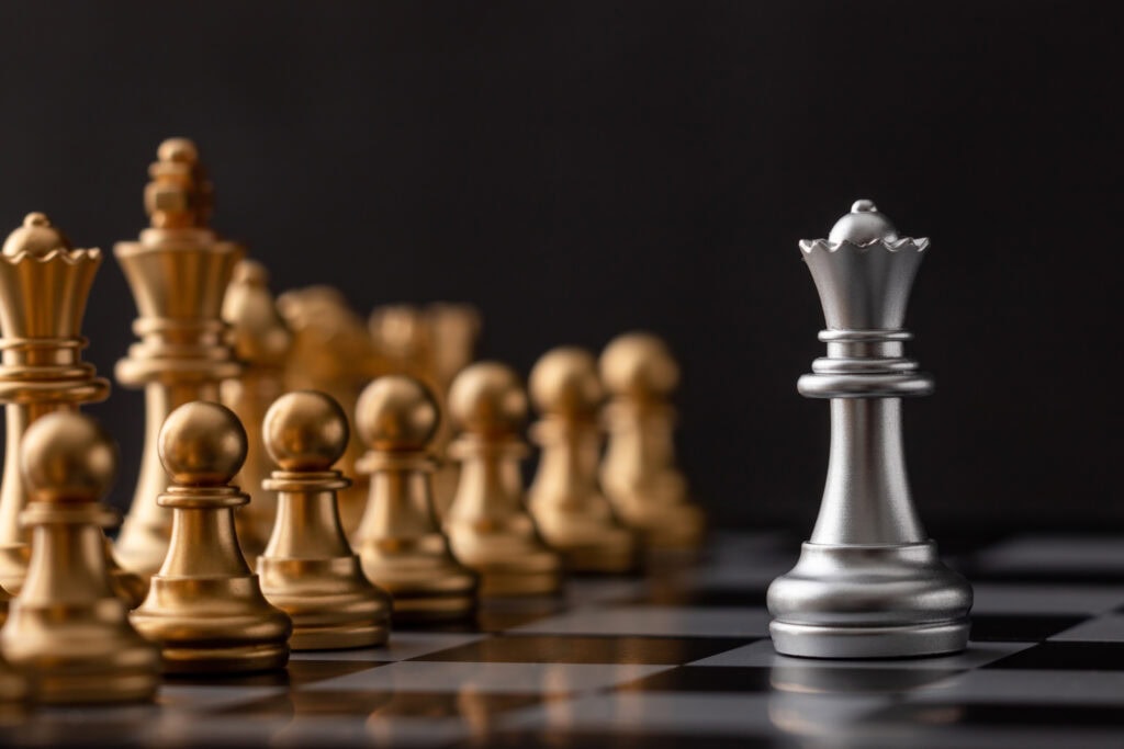 A silver queen chess piece stands apart from a row of gold chess pieces on a black and white chessboard with a dark background.