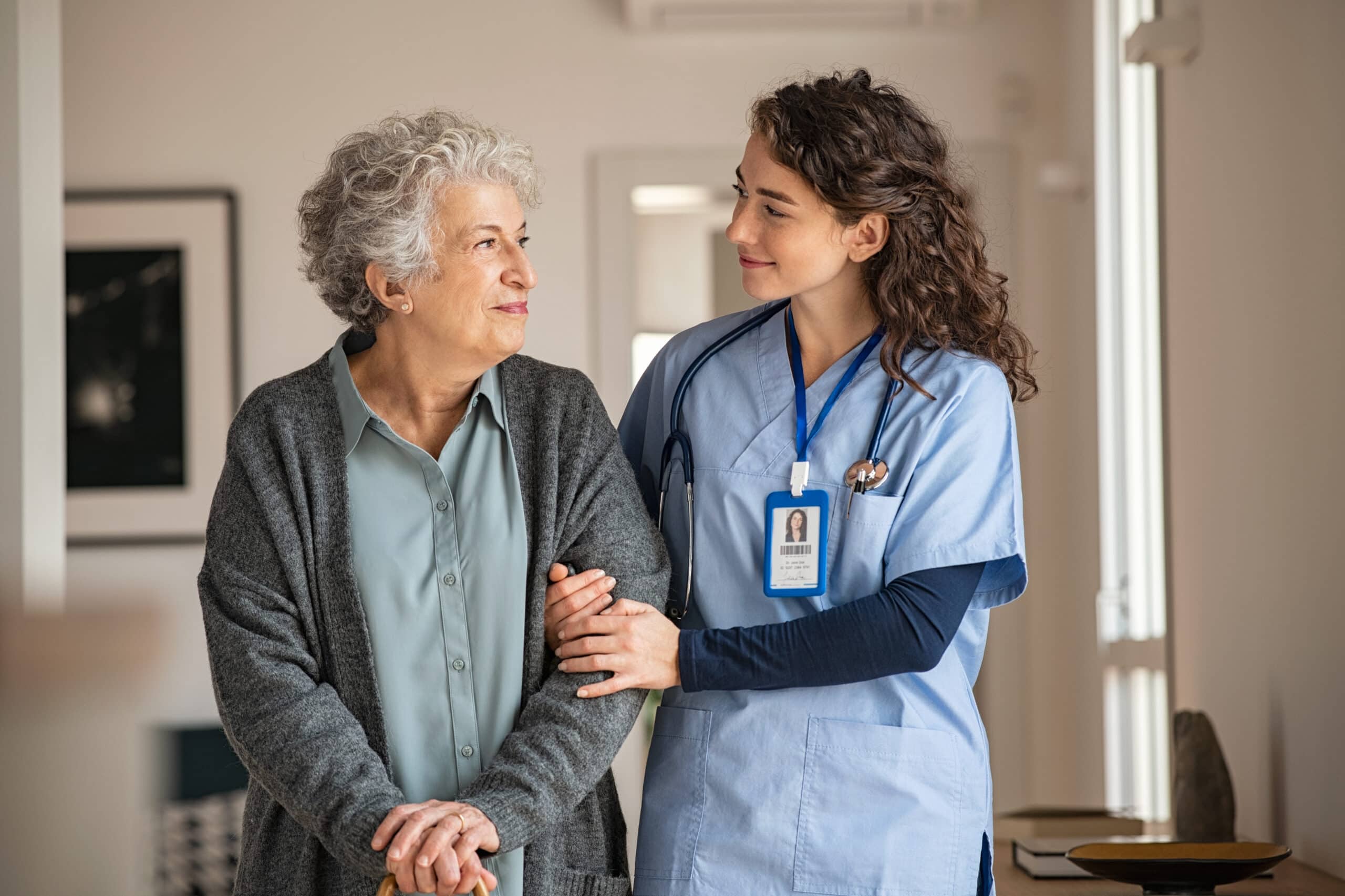 An elderly woman stands with a cane next to a healthcare worker in scrubs, who is smiling and holding her arm in a supportive manner.