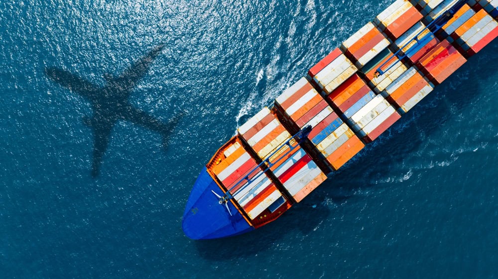 Aerial view of a large cargo ship carrying colorful containers at sea, with the shadow of an airplane visible on the water.