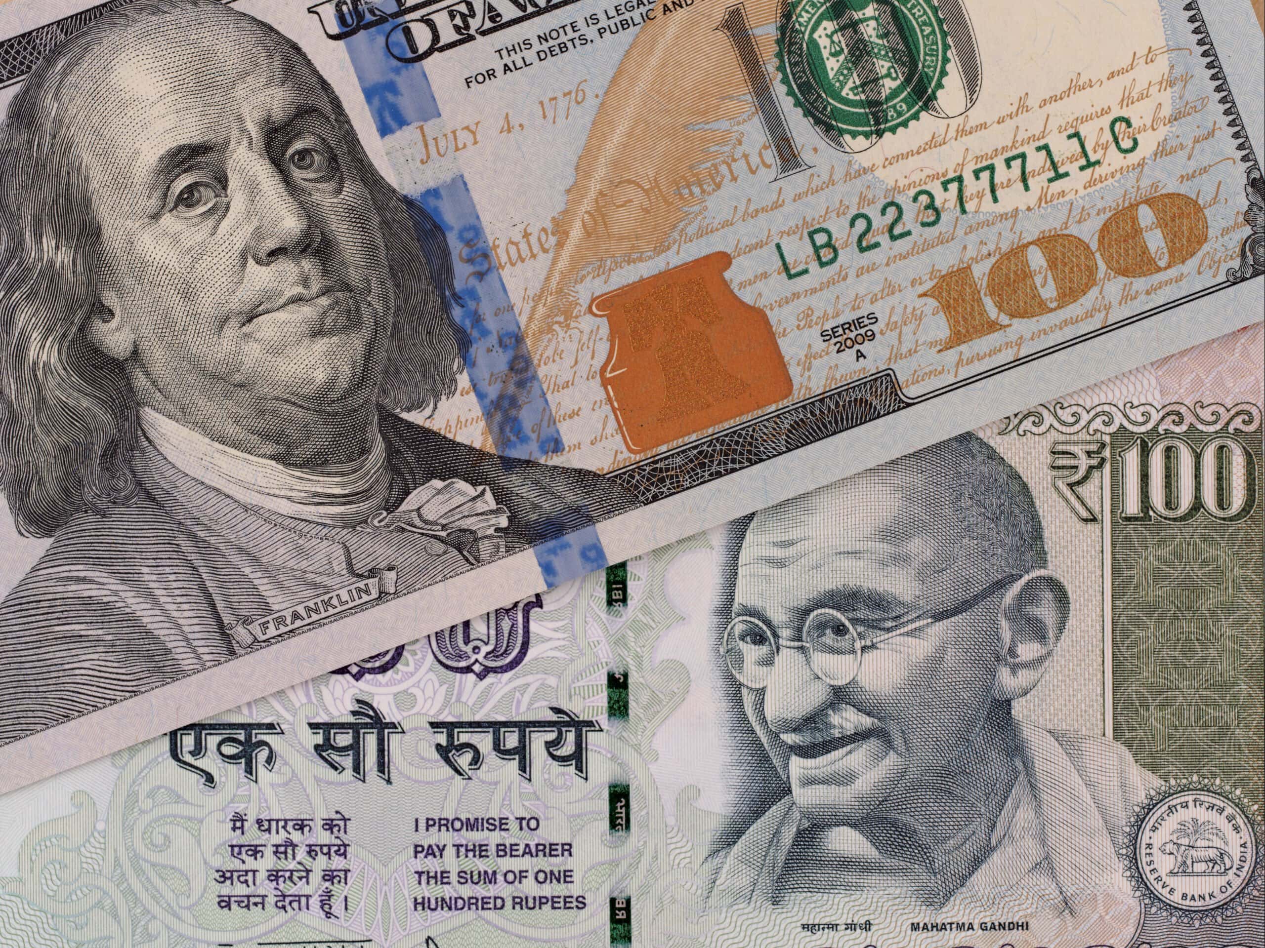A close-up of a US 100 dollar bill featuring Benjamin Franklin and an Indian 100 rupee note featuring Mahatma Gandhi placed side by side.