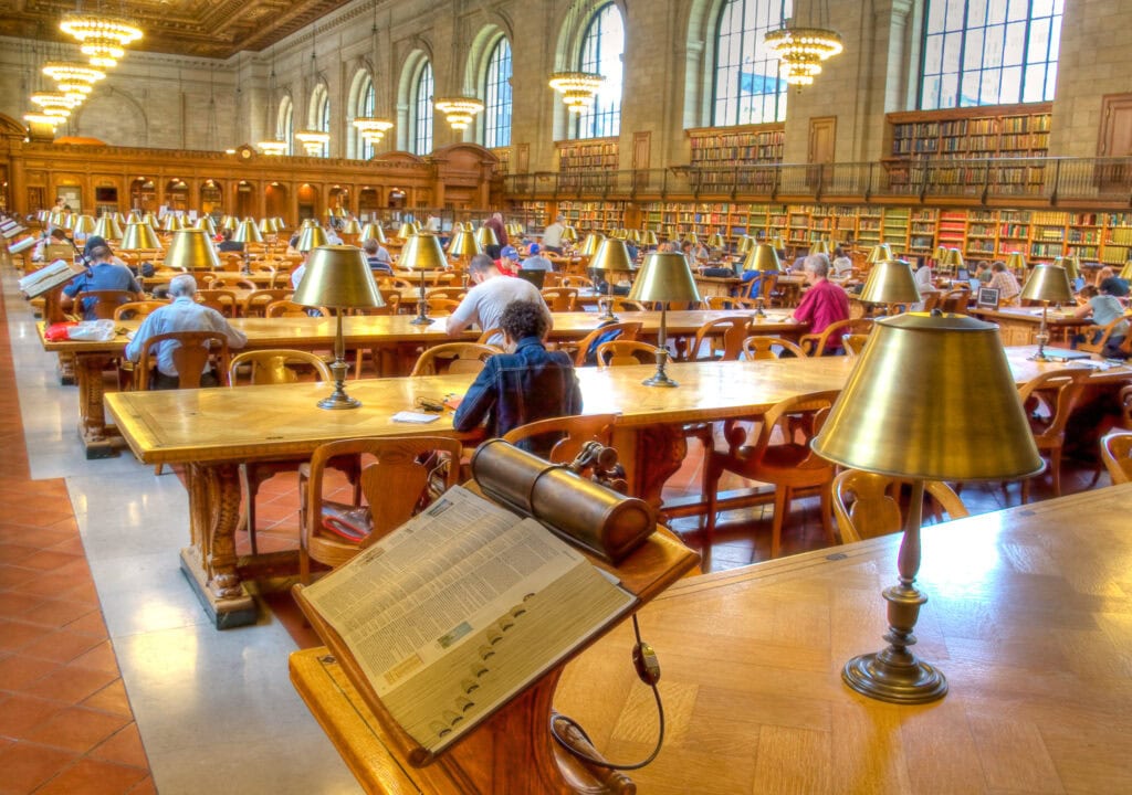 People sit and read at long wooden tables in a large, ornate library room with tall windows, chandeliers, and shelves filled with books.