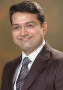 A man wearing glasses, a dark pinstriped suit, a white shirt, and a patterned tie, smiling in front of a plain brown background.