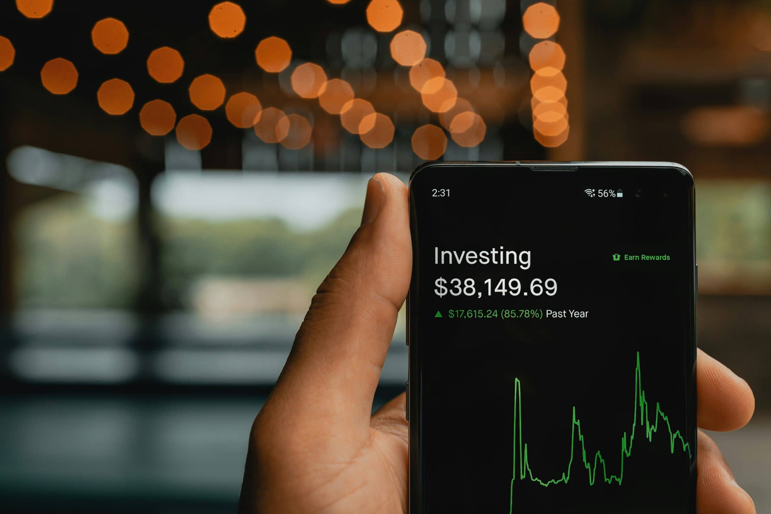 A hand holds a smartphone displaying an investing app with a portfolio value of $38,149.69, showing a green upward trend graph and blurred lights in the background.