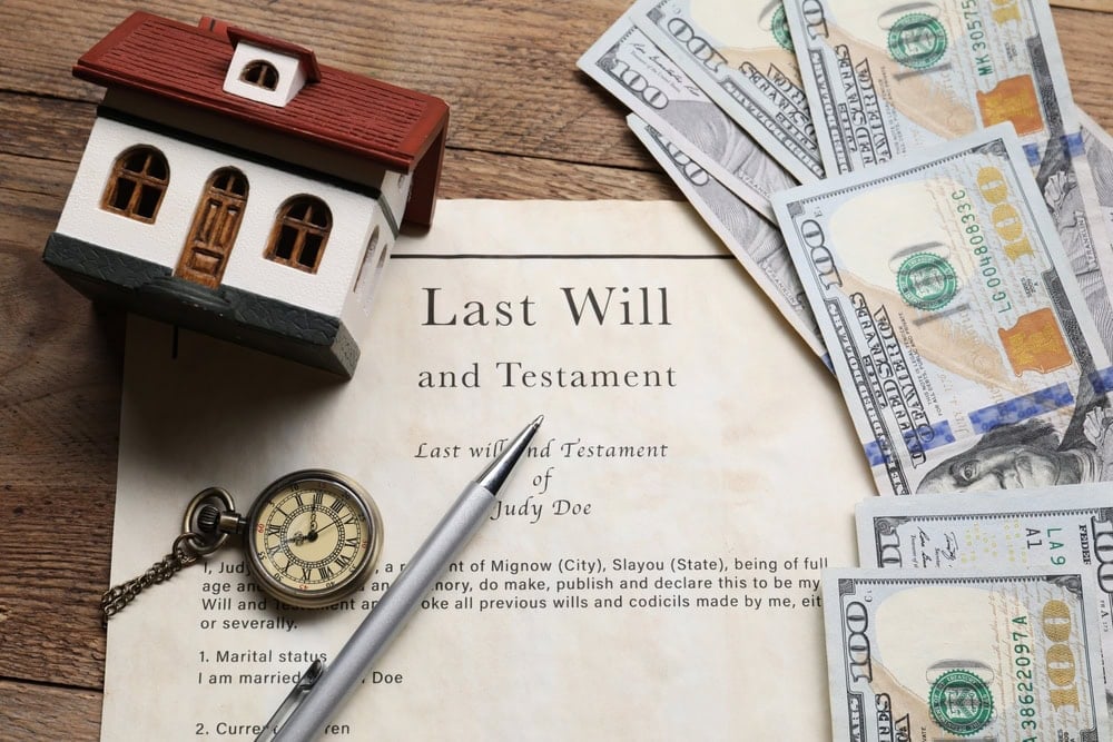 A last will and testament document on a table with a pen, a small house model, a pocket watch, and several hundred-dollar bills.
