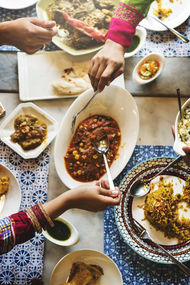 Several people serve themselves from shared dishes of colorful Indian food at a dining table, with rice, curries, and condiments arranged on patterned tablecloths.