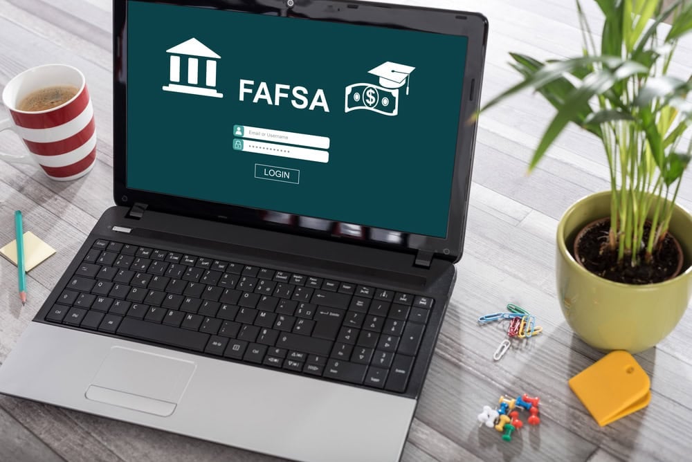 A laptop displays a FAFSA login screen on a desk with a coffee cup, plant, paper clips, and a yellow keychain nearby.
