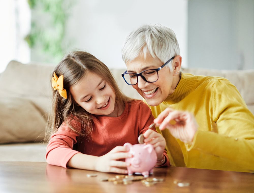 An older woman and a young girl smiling while putting coins into a pink piggy bank at a table.