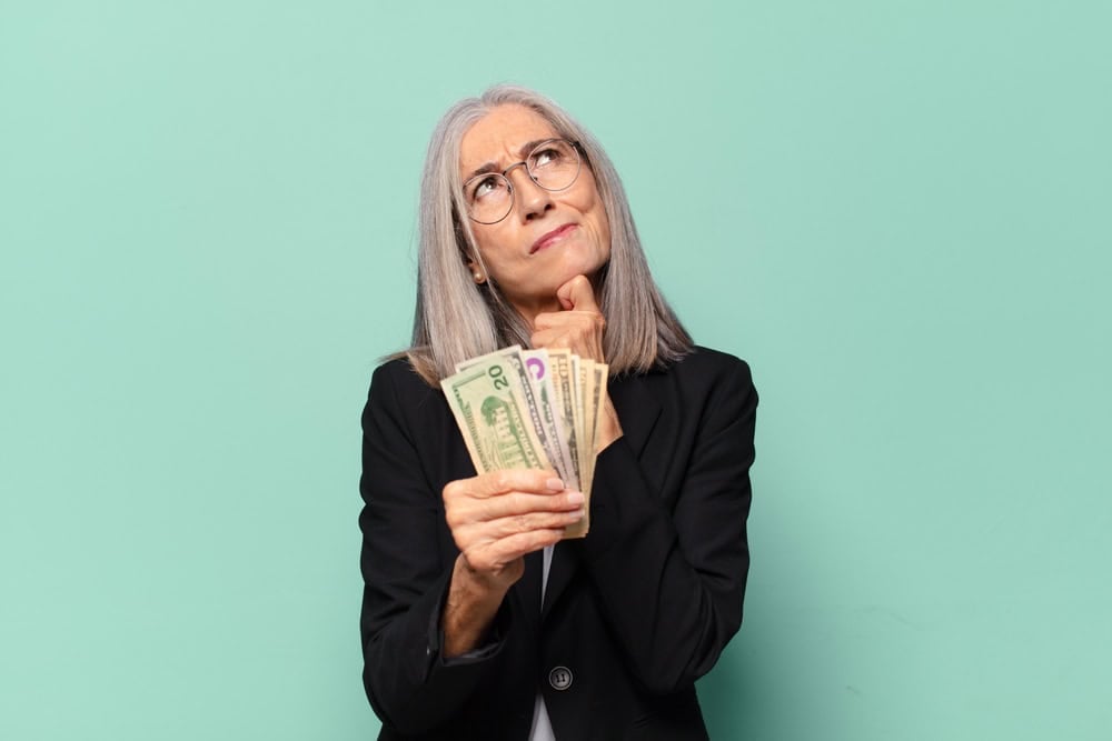 Older woman in a blazer holds a stack of U.S. dollar bills and looks upward with a thoughtful expression against a mint green background.