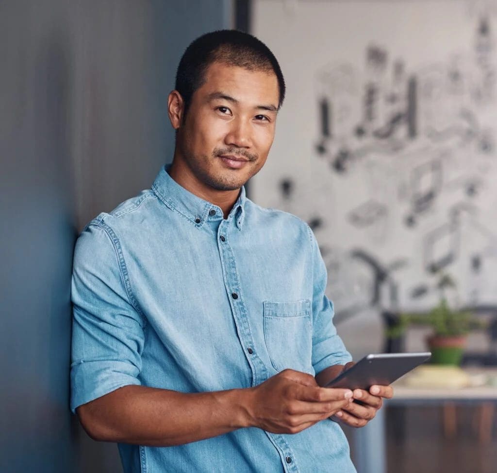 A man in a light blue denim shirt stands indoors, holding a tablet and looking at the camera, with a blurred background featuring a wall mural and a potted plant.