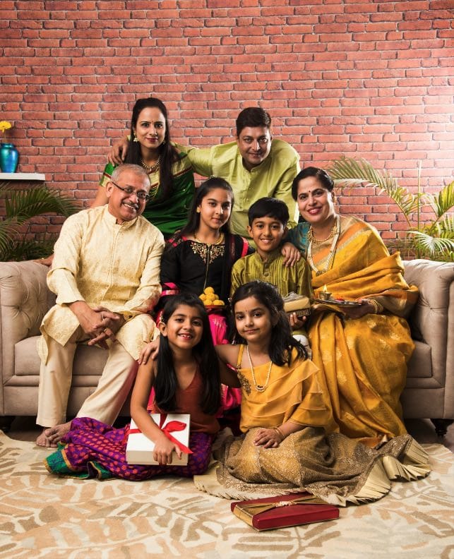 A multi-generational family in traditional Indian attire poses together on a sofa with gifts and sweets, against a brick wall and indoor plants.