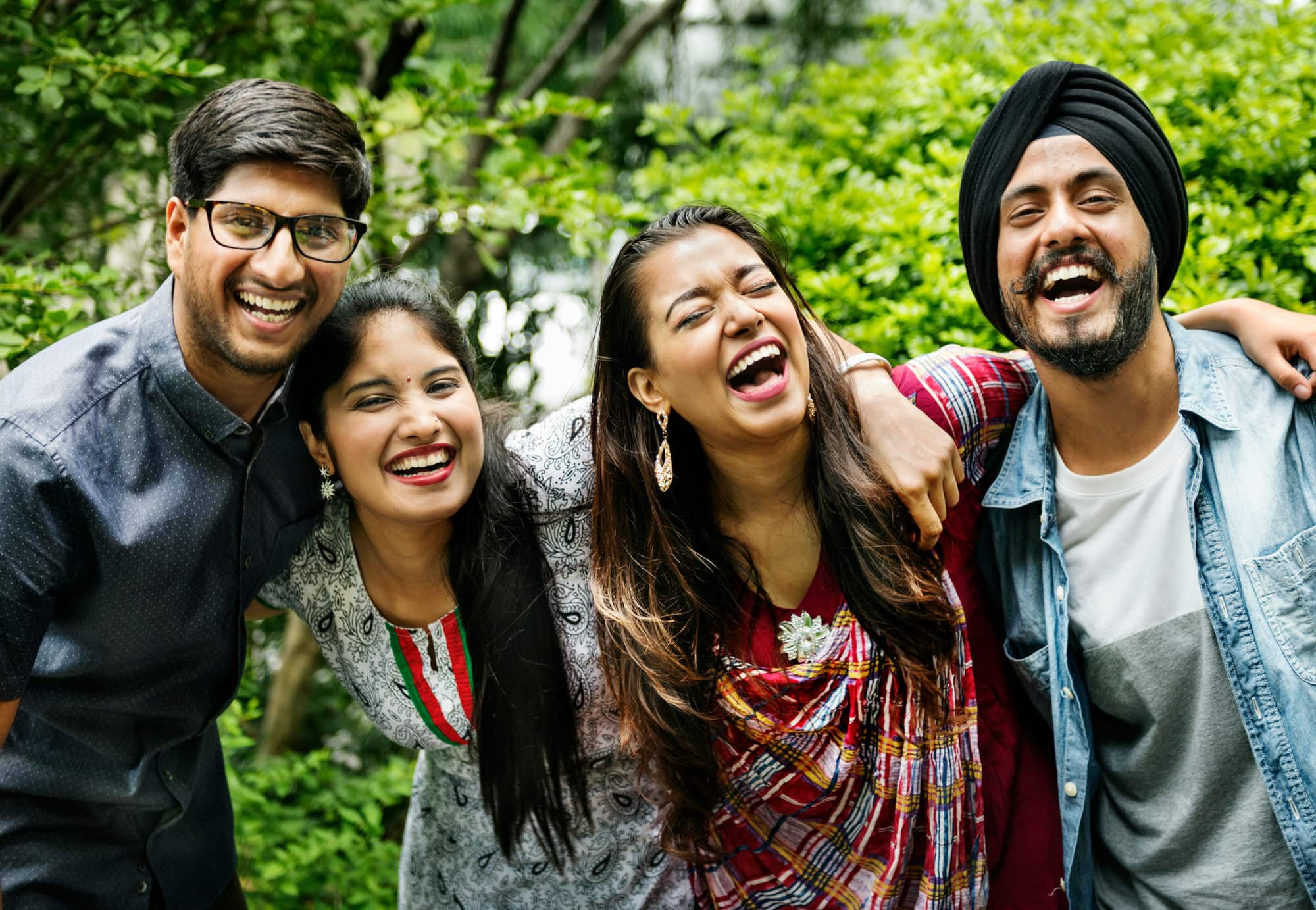 Four people stand close together outdoors, smiling and laughing, with greenery in the background.