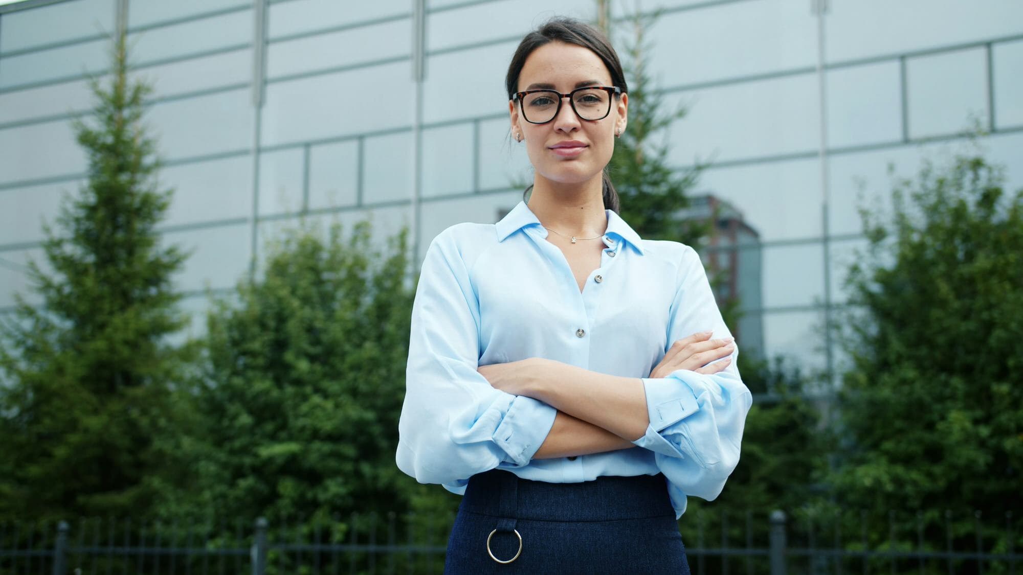 A woman wearing glasses and a light blue shirt stands outdoors with arms crossed, in front of a building and trees.