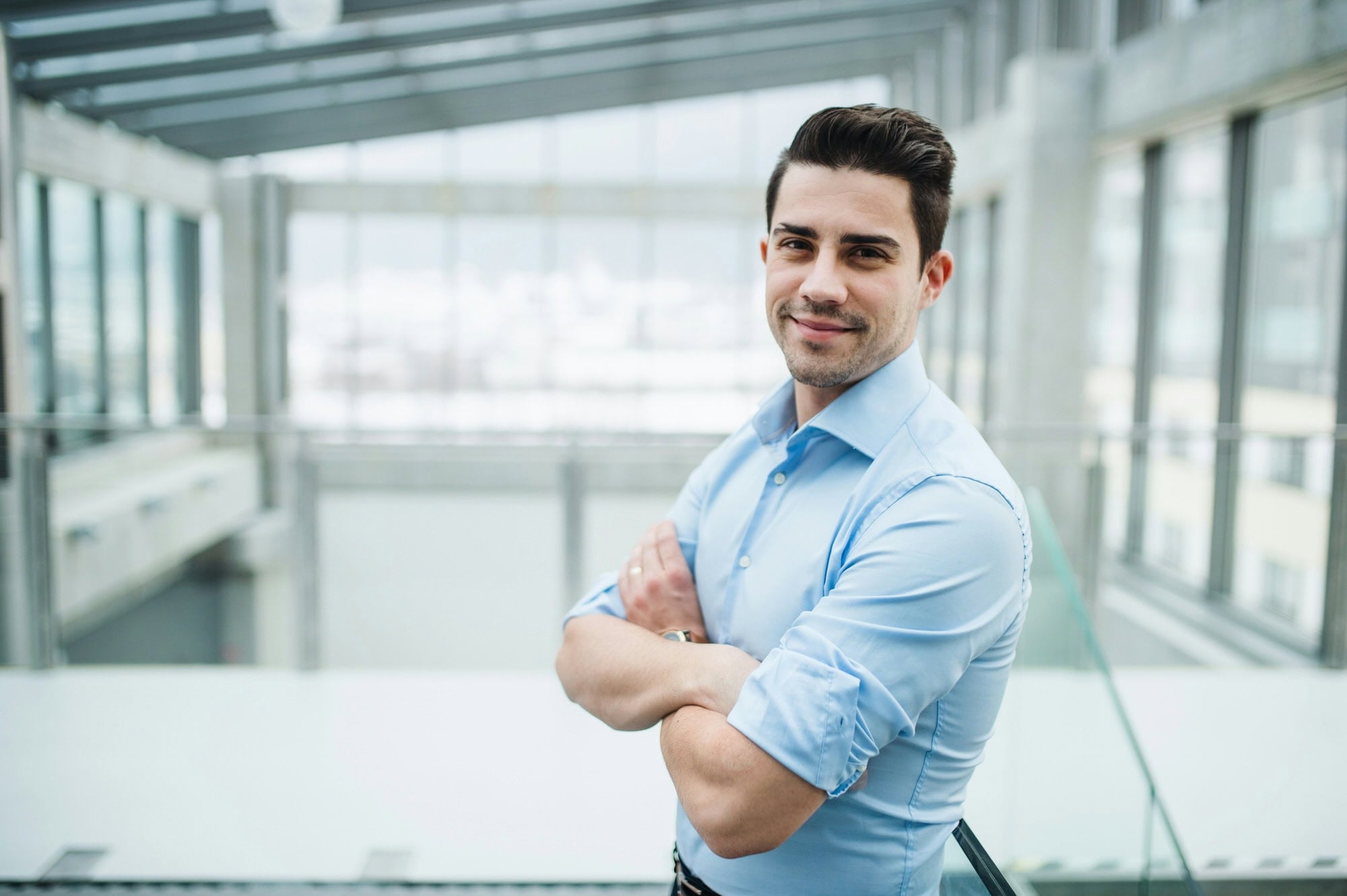 A man in a light blue shirt stands indoors with arms crossed, smiling at the camera. The background shows large windows and modern architecture.