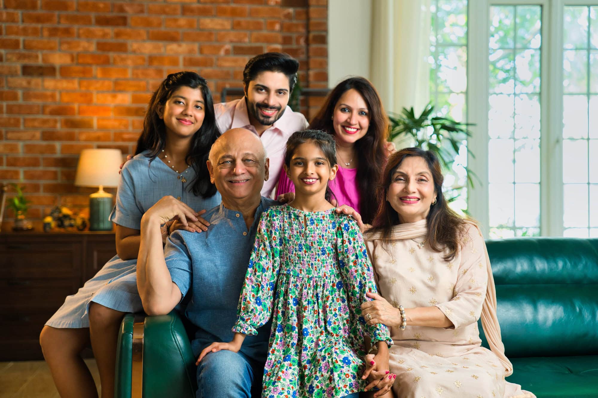 Three generations of a family, including two children, their parents, and grandparents, sit together on a couch in a well-lit living room, smiling at the camera.