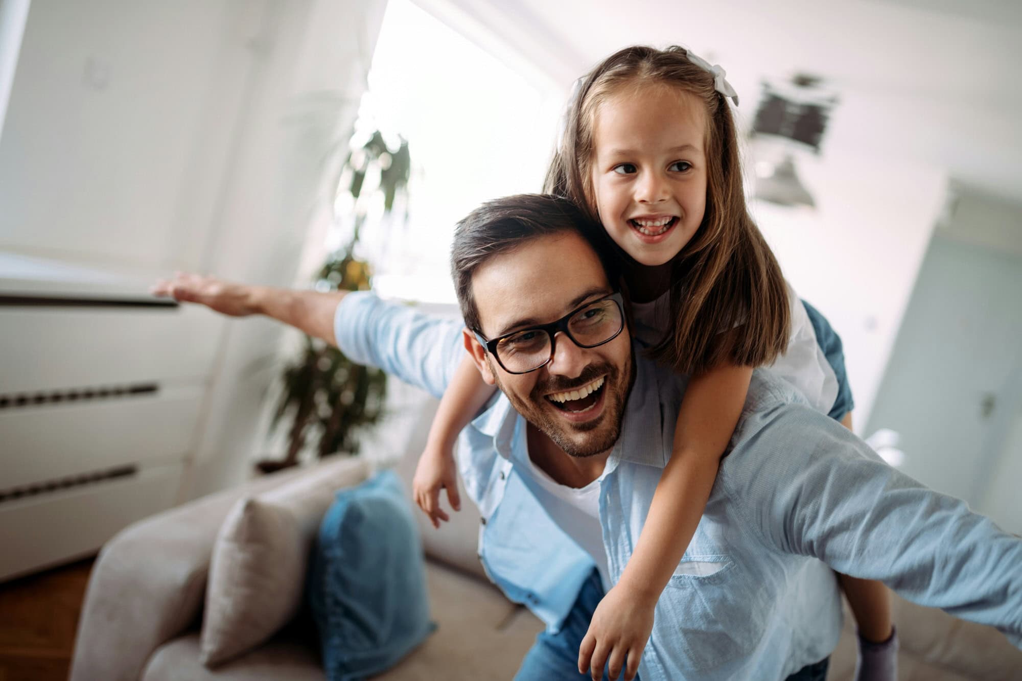 A man with glasses smiles while giving a piggyback ride to a young girl indoors; both have their arms outstretched and appear happy.