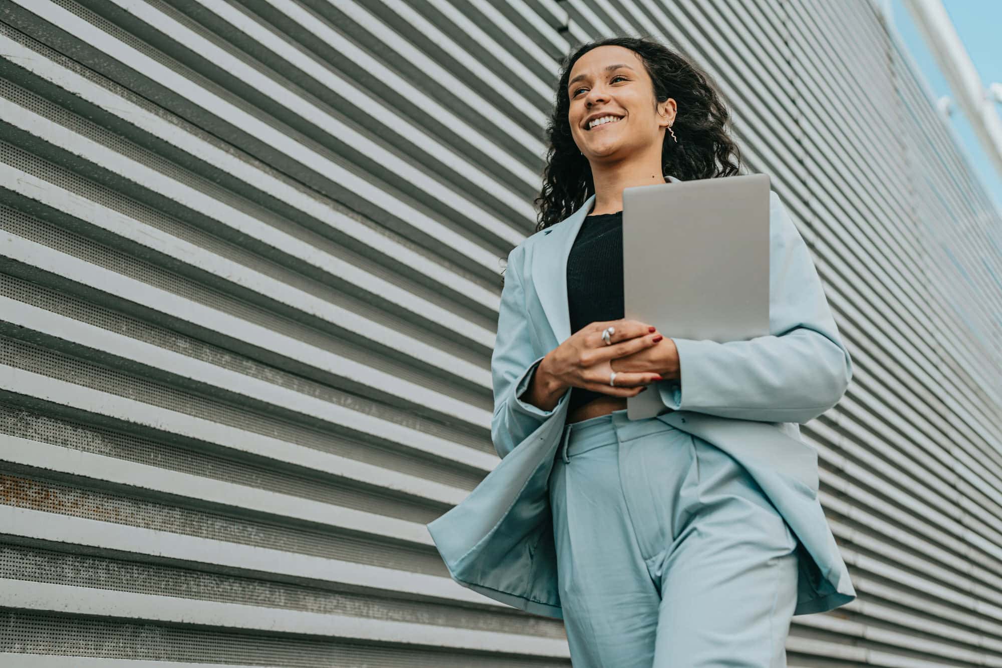 A person in a light blue suit walks outdoors carrying a closed laptop, smiling, with a corrugated metal wall in the background.