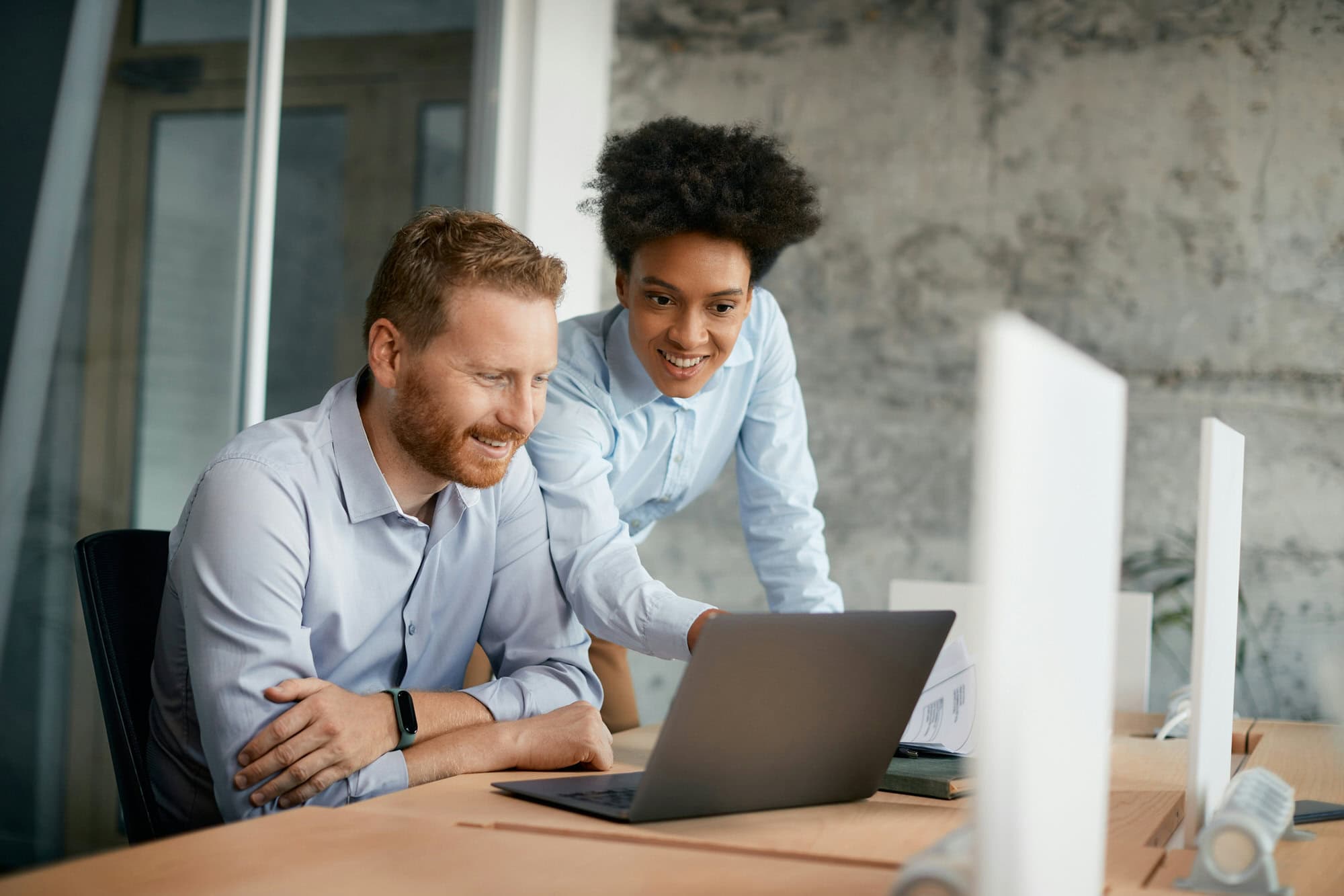 Two colleagues in a modern office look at a laptop screen together, one sitting and smiling while the other stands and gestures at the screen.