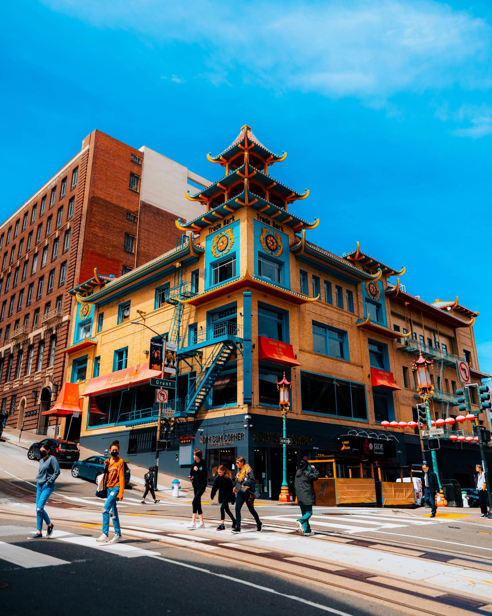 People cross a street at an intersection in front of a colorful building with traditional East Asian architectural design on a sunny day in an urban area.