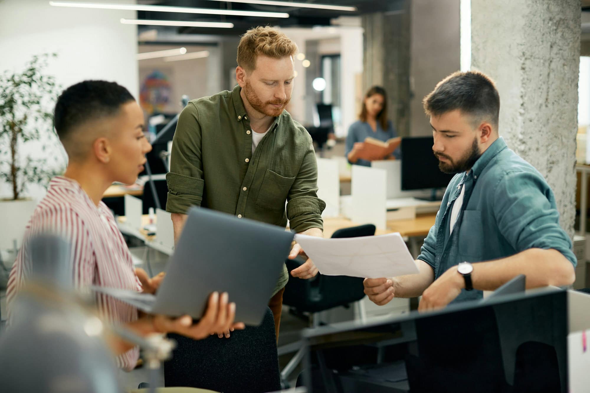 Three people in a modern office discuss documents and a laptop while standing by a desk, with another person working in the background.