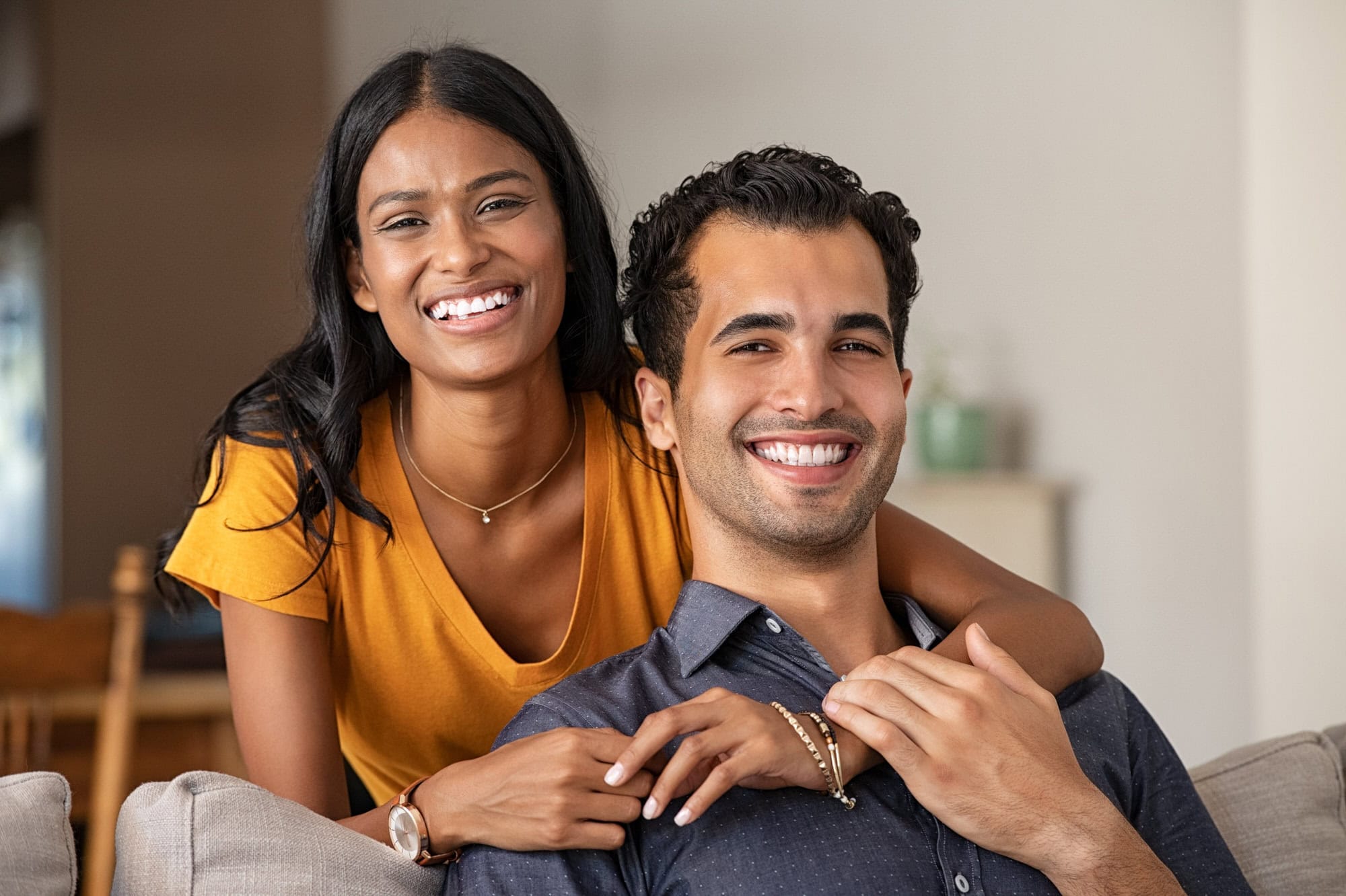 A woman in a yellow top stands behind a man in a blue shirt as they both sit on a couch and smile at the camera.