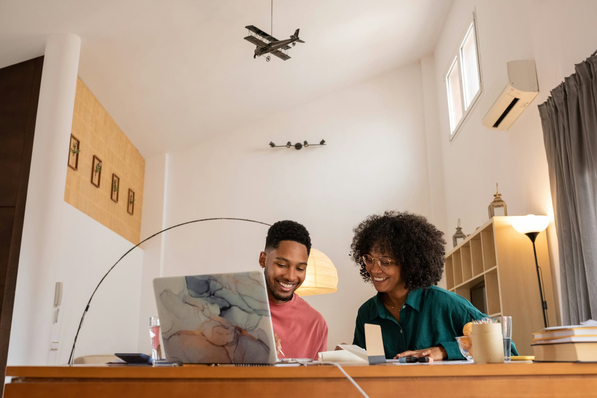 Two people sit at a desk working on a laptop together in a bright, modern room with model airplanes hanging from the ceiling.