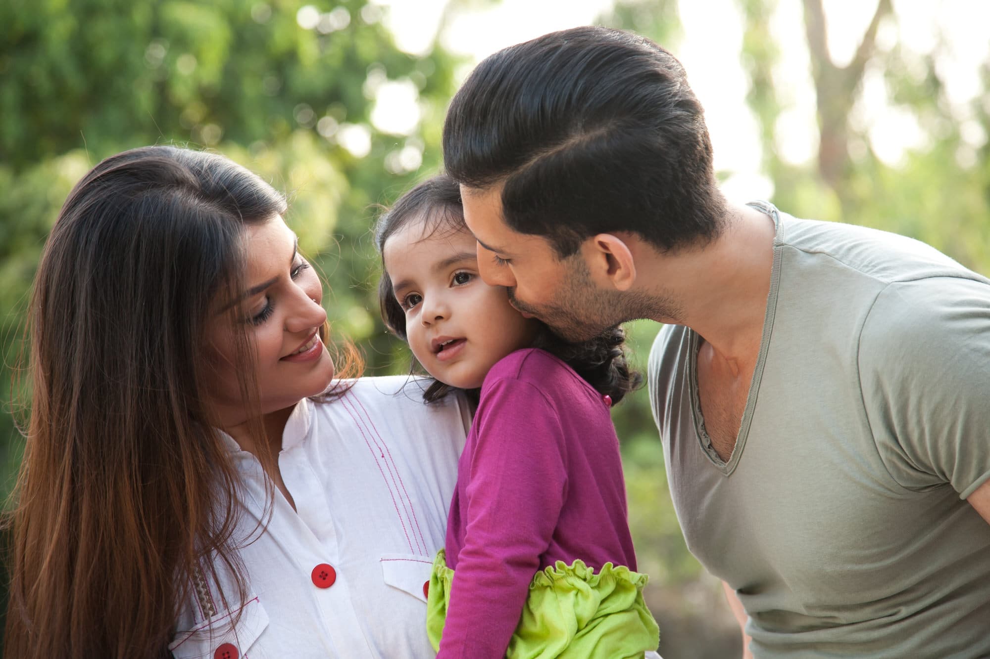 A man kisses a young girl on the cheek while a woman smiles at them; they are outdoors with green foliage in the background.