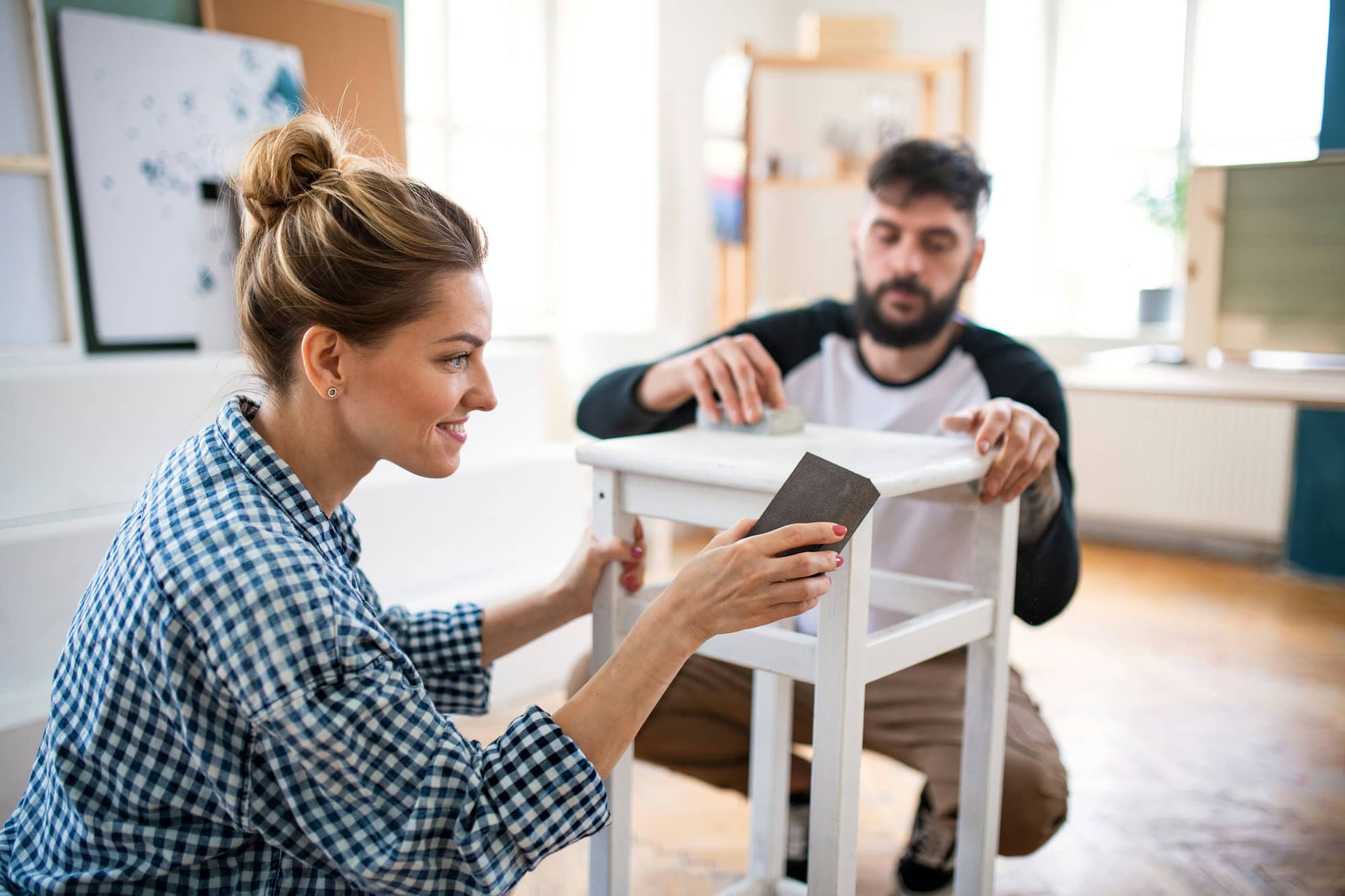 A woman and a man are sanding a white wooden stool together in a bright, indoor room. The woman is using a sanding block, and both appear focused on the task.