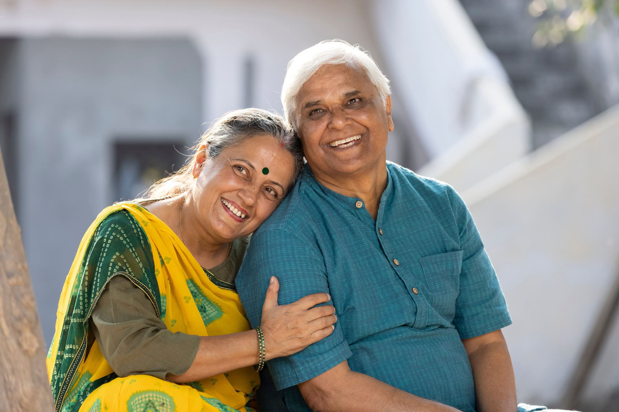 An older woman in a yellow sari sits outdoors, smiling and leaning her head on the shoulder of an older man in a blue kurta, who is also smiling.