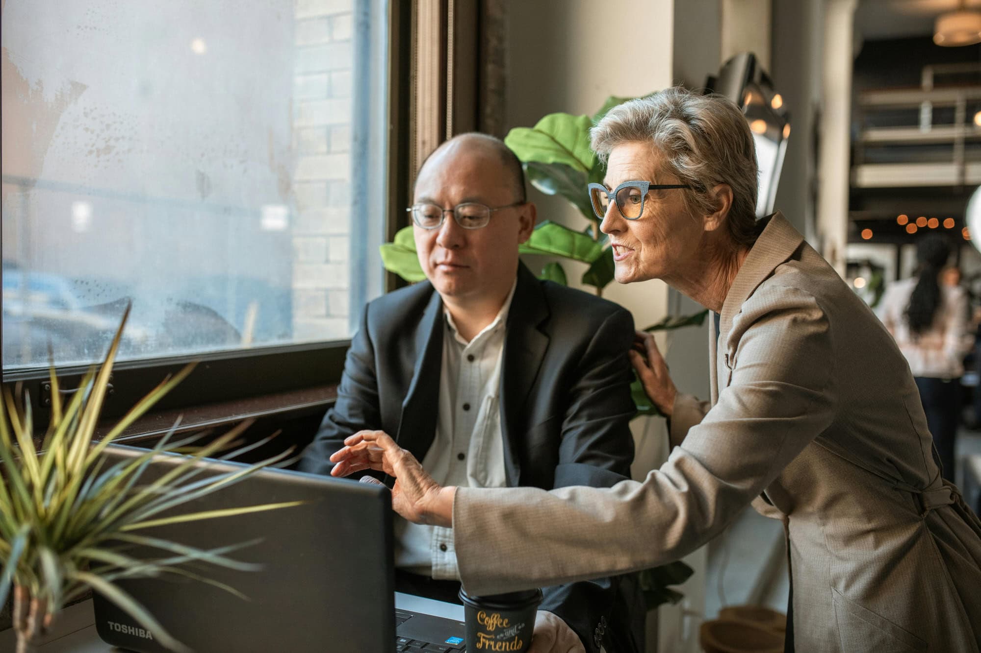 Two professionally dressed adults work together at a laptop by a window; one is seated and looking at the screen while the other points at it and speaks.