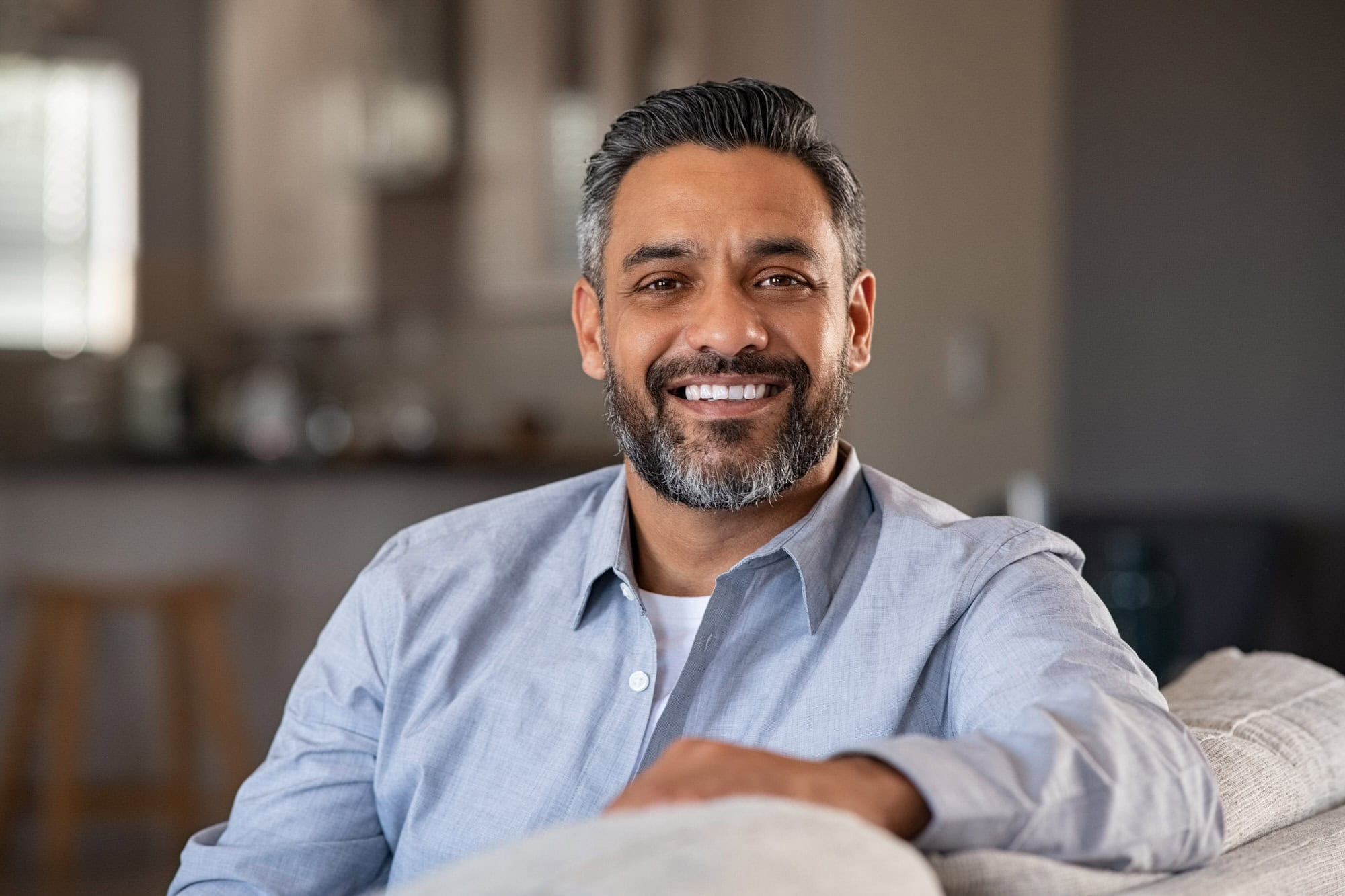 A man with dark hair and a beard, wearing a light blue shirt, sits on a couch and smiles at the camera in a home setting.