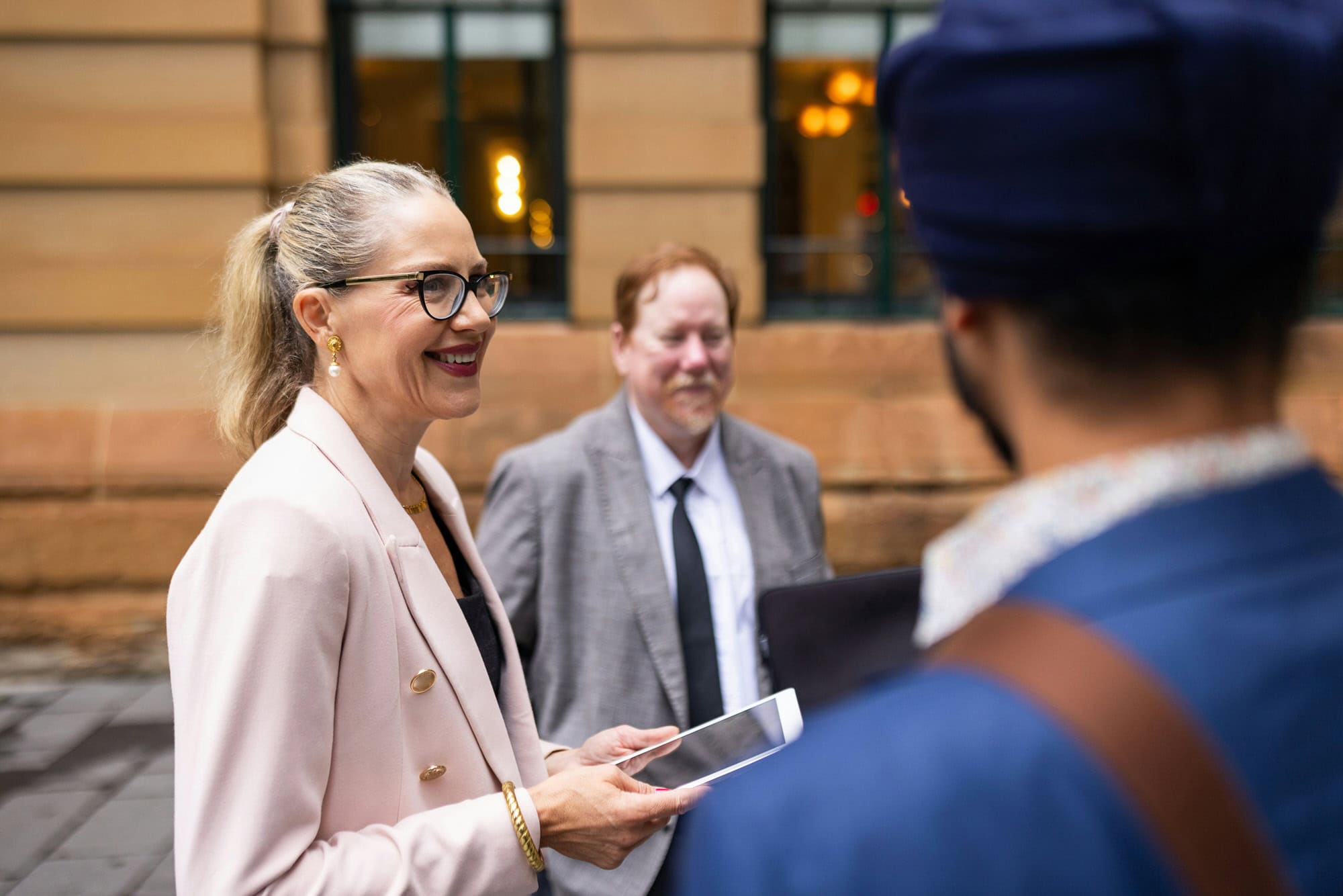 A woman in a light blazer holds a tablet and smiles while talking to two men outside a building.