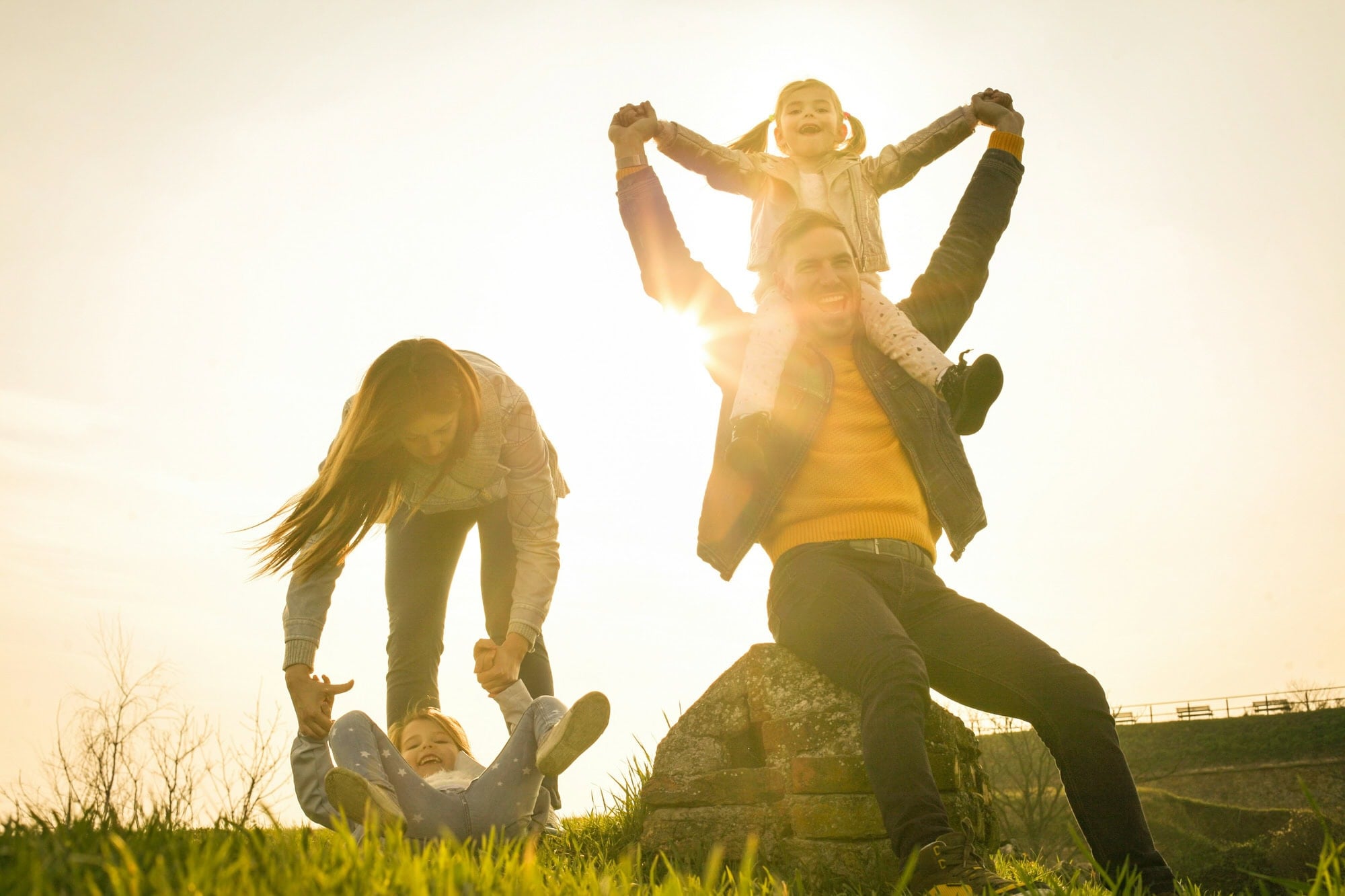 Two adults and two children play outdoors at sunset; one child sits on an adult’s shoulders while the other is swung by the second adult.