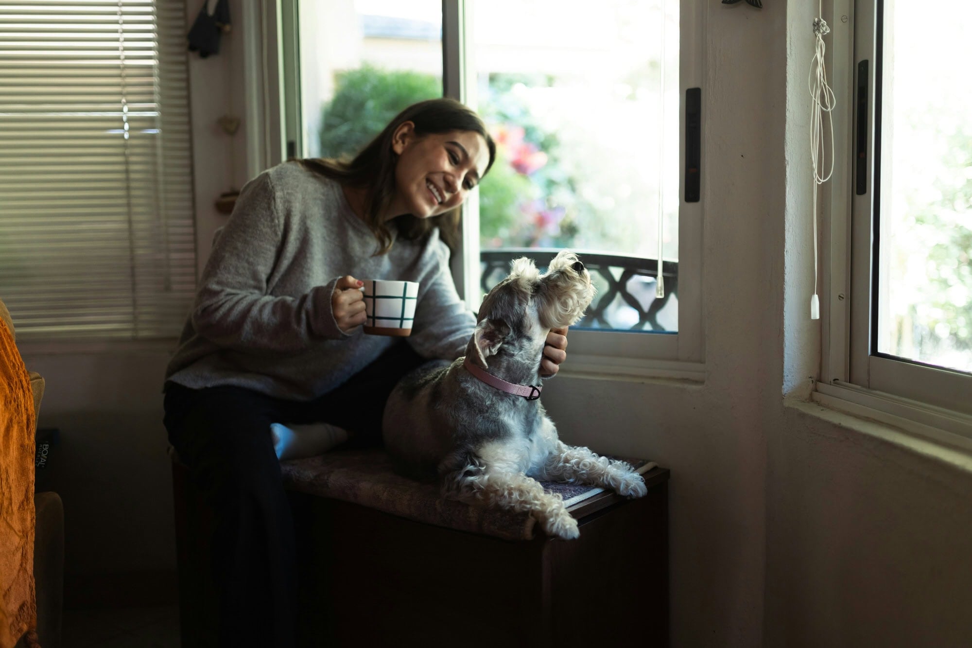 A woman sits by a window holding a mug, smiling and petting a small dog that is looking up at her.