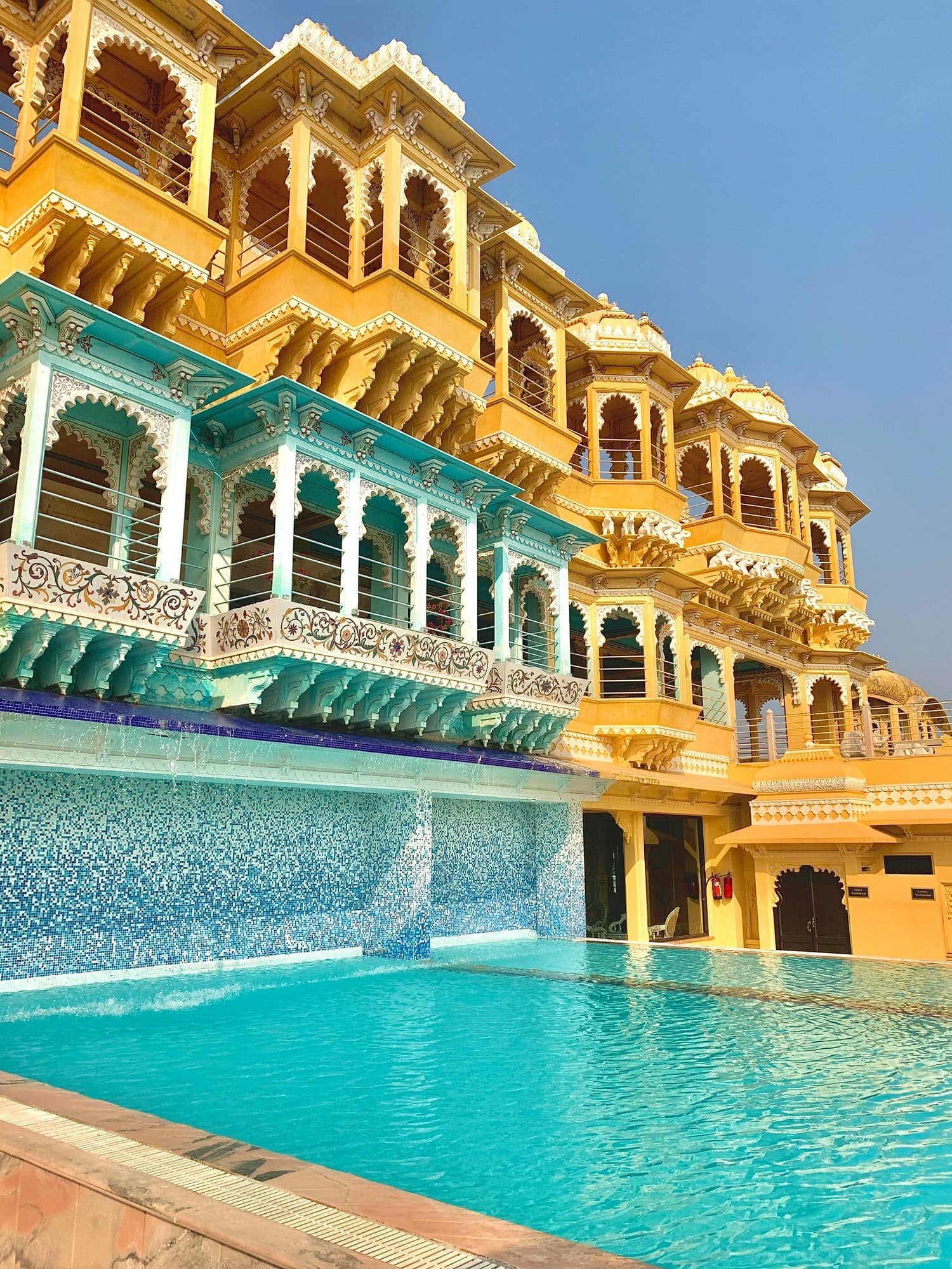 A traditional building with ornate yellow and blue facades stands beside a clear outdoor pool under a bright blue sky.