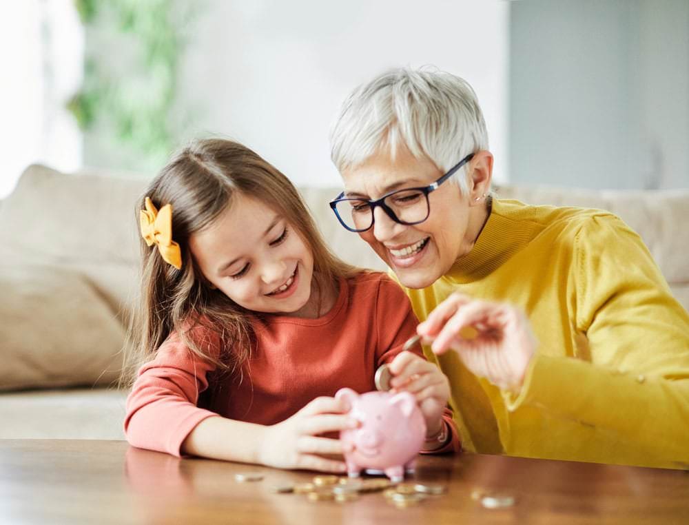 An older woman and a young girl smile as they put coins into a pink piggy bank on a table at home.