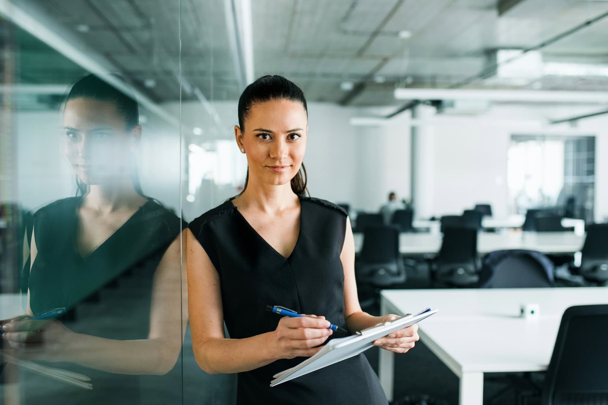 Woman standing in a modern office, holding a clipboard and pen, looking at the camera with empty desks and computers in the background.