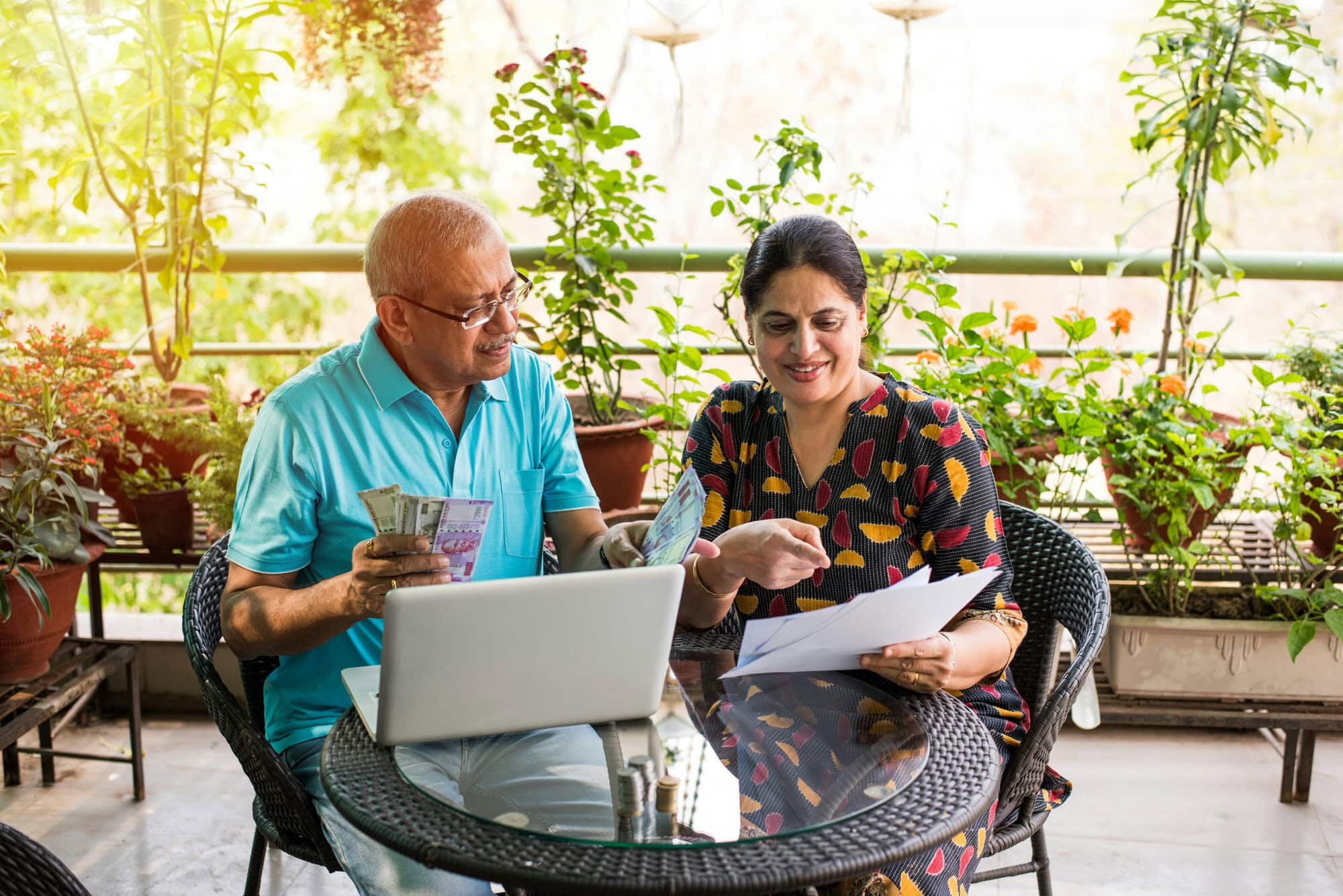 An older man and woman sit at a table with a laptop, holding cash and documents, surrounded by potted plants on a balcony.