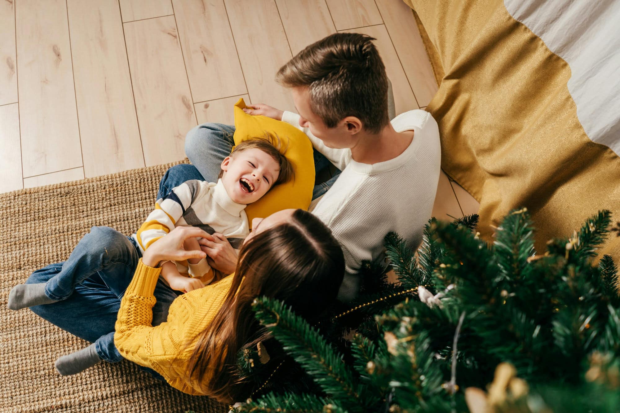 An adult man and woman sit on the floor with a young boy, who is laughing. They are near a Christmas tree and a yellow blanket.