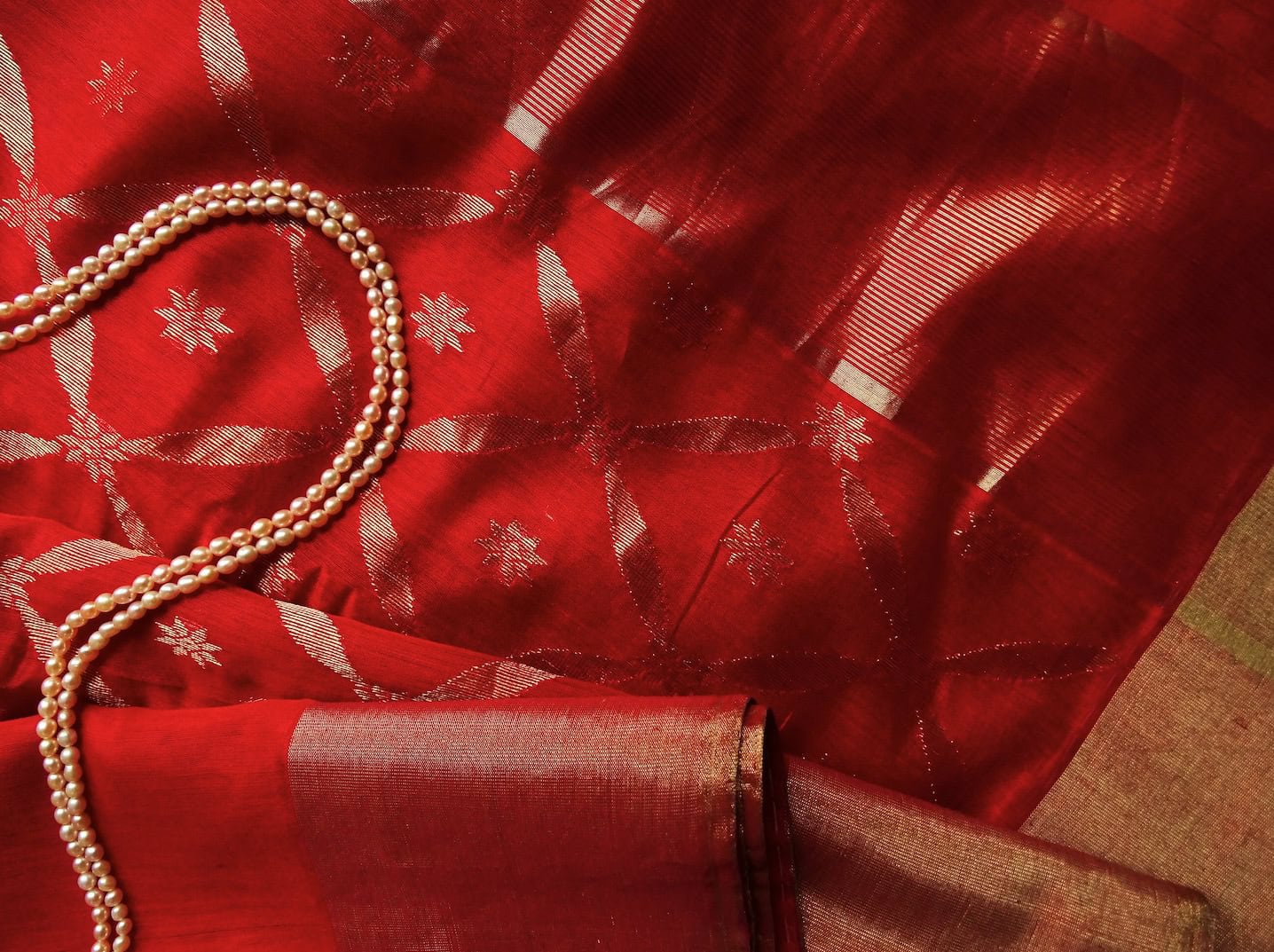 A red silk saree with golden floral patterns is displayed alongside a string of pearls.