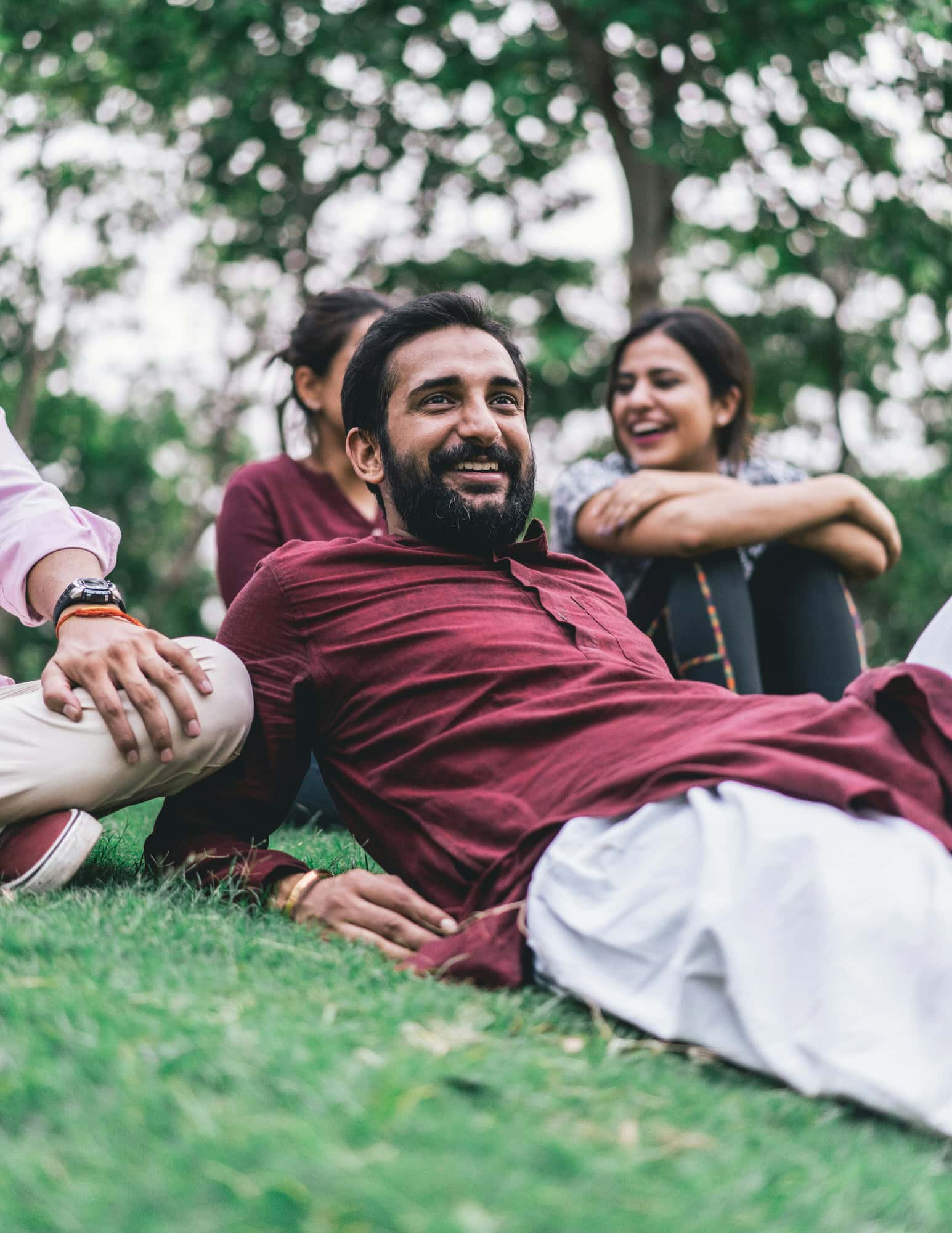 A man in a maroon kurta sits on the grass with others, smiling and relaxing outdoors with trees in the background.
