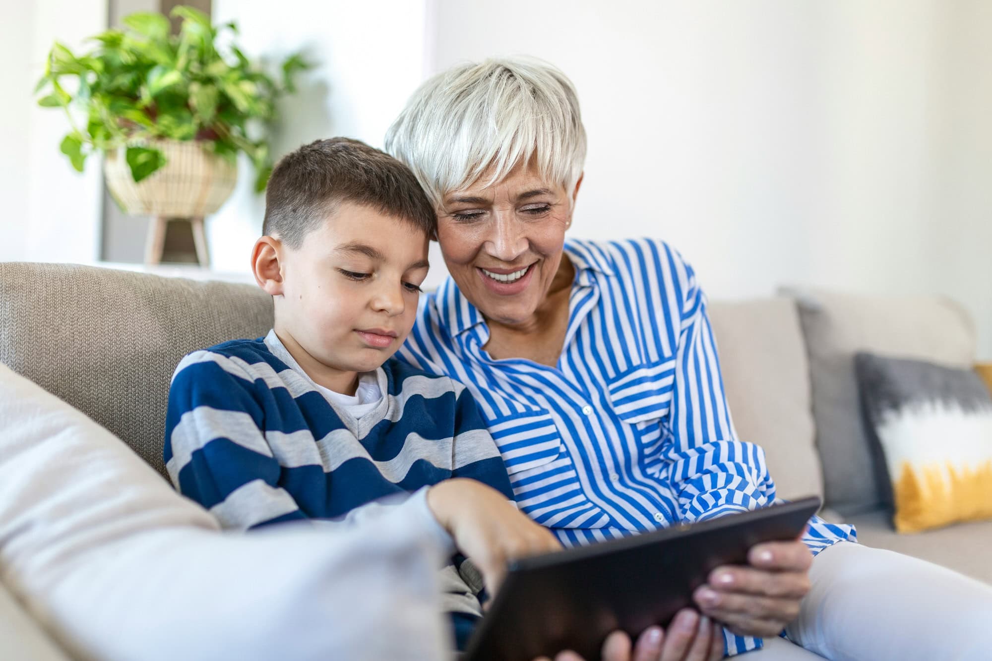 An older woman and a young boy sit on a couch, looking at a tablet together. The boy points at the screen while the woman smiles.