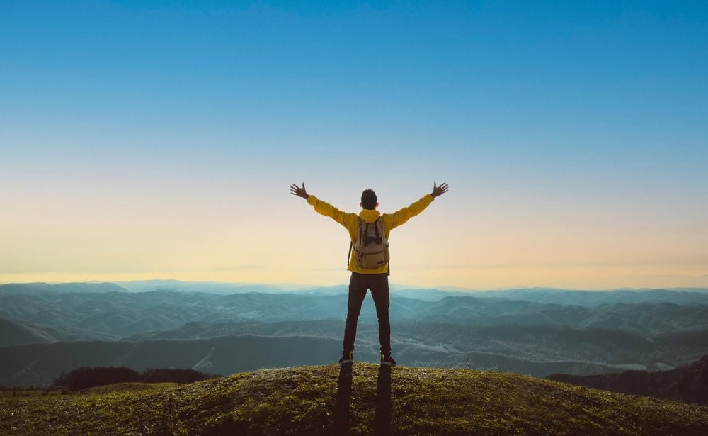 A person wearing a yellow jacket and backpack stands on a hilltop with arms outstretched, overlooking a vast landscape of mountains under a clear sky.