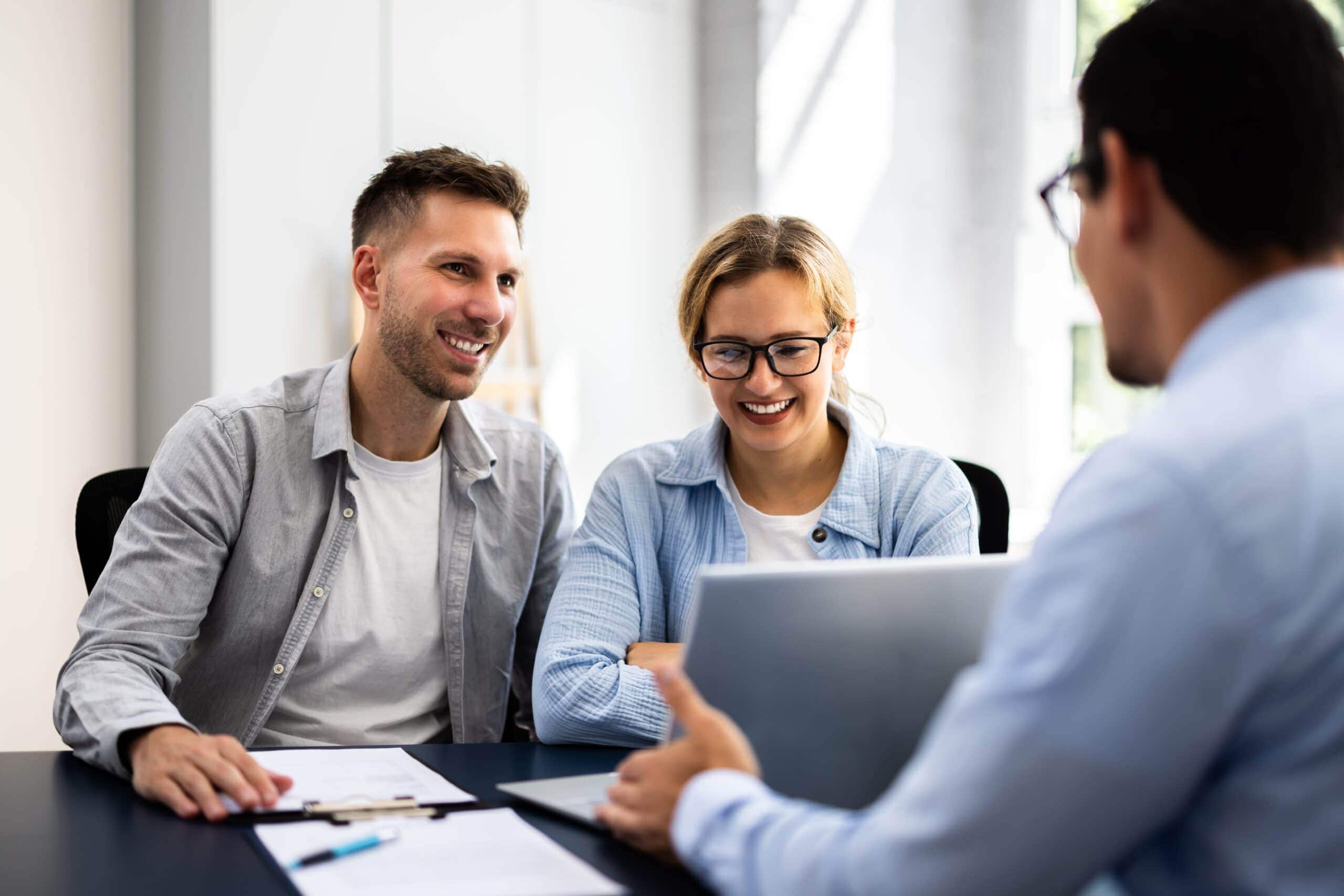 A man and woman sit at a desk with paperwork, smiling and listening to another person who is showing them something on a laptop.