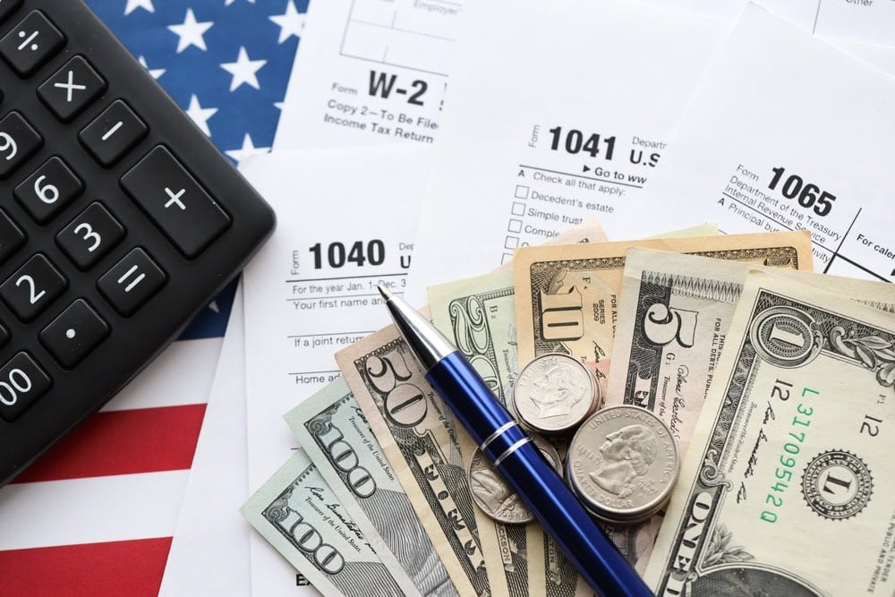 A calculator, pen, US dollar bills and coins, and various IRS tax forms are displayed on top of an American flag.