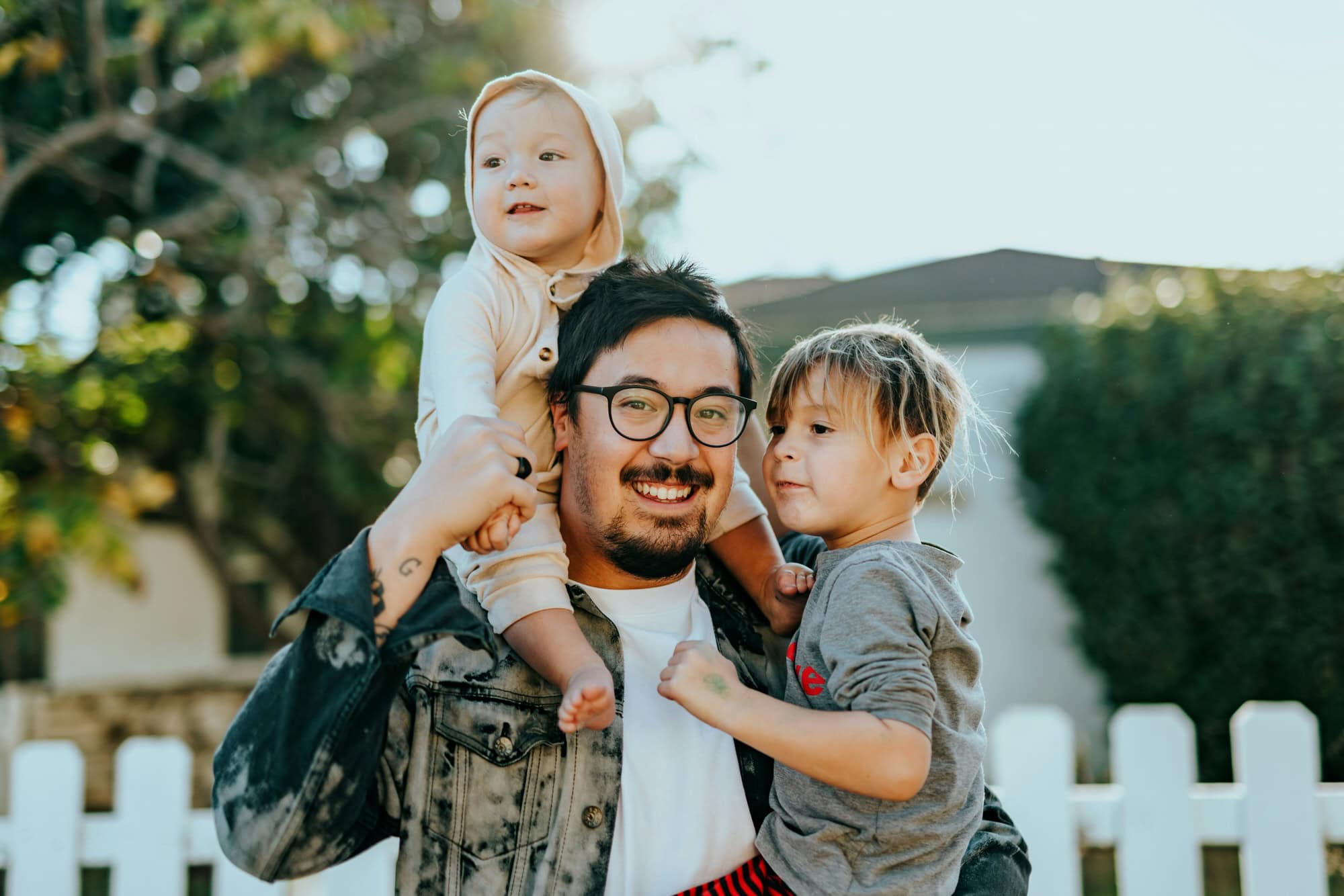 A man wearing glasses smiles while holding a baby on his shoulder and a young child in his arm, standing outside near a white picket fence.
