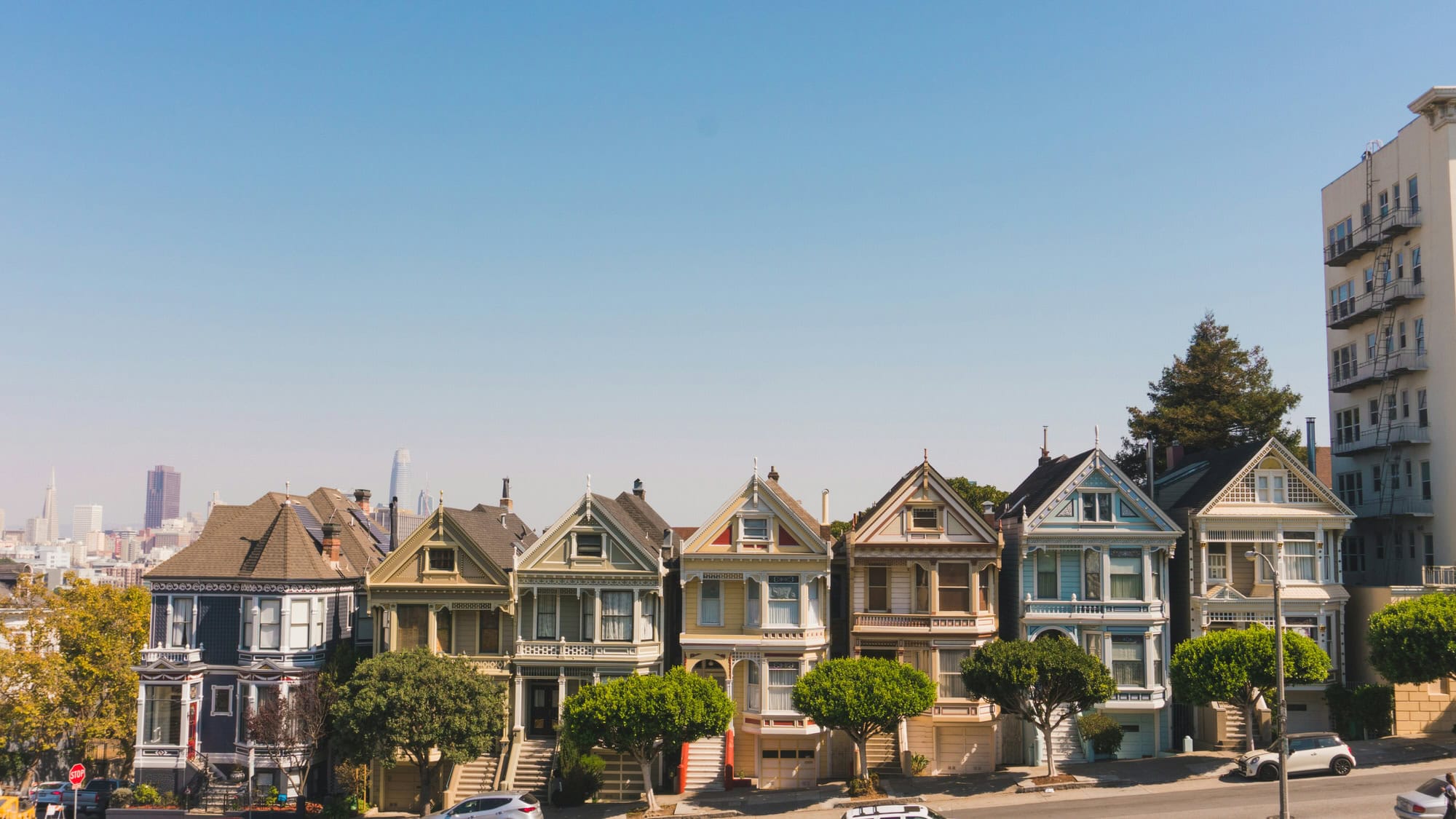 A row of colorful Victorian houses, known as the Painted Ladies, stands in front of a clear blue sky in San Francisco, with modern buildings visible in the background.